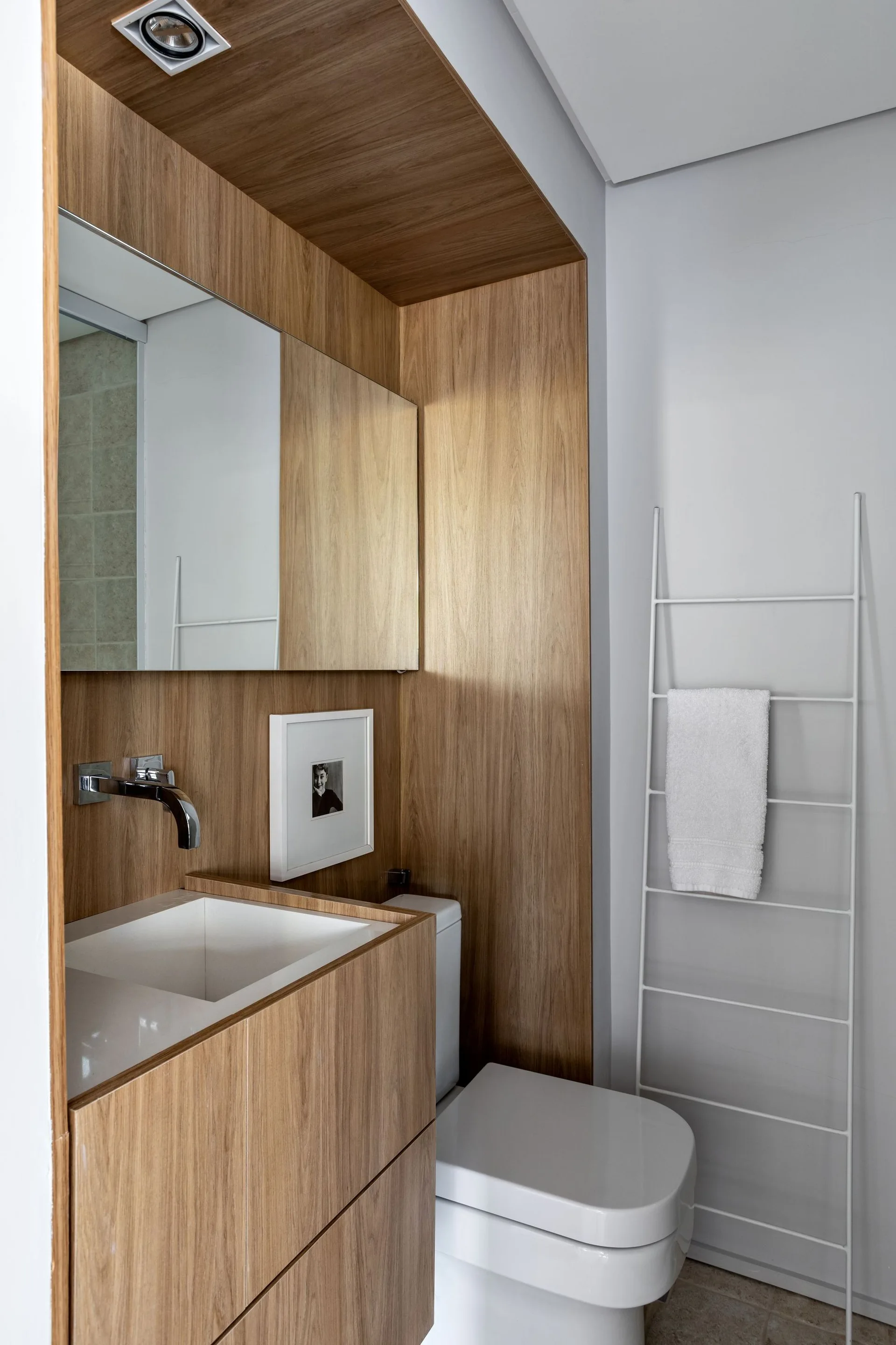 A modern powder room features a wood-paneled vanity niche, a white undermount sink with a wall-mounted faucet, and a leaning towel ladder.
