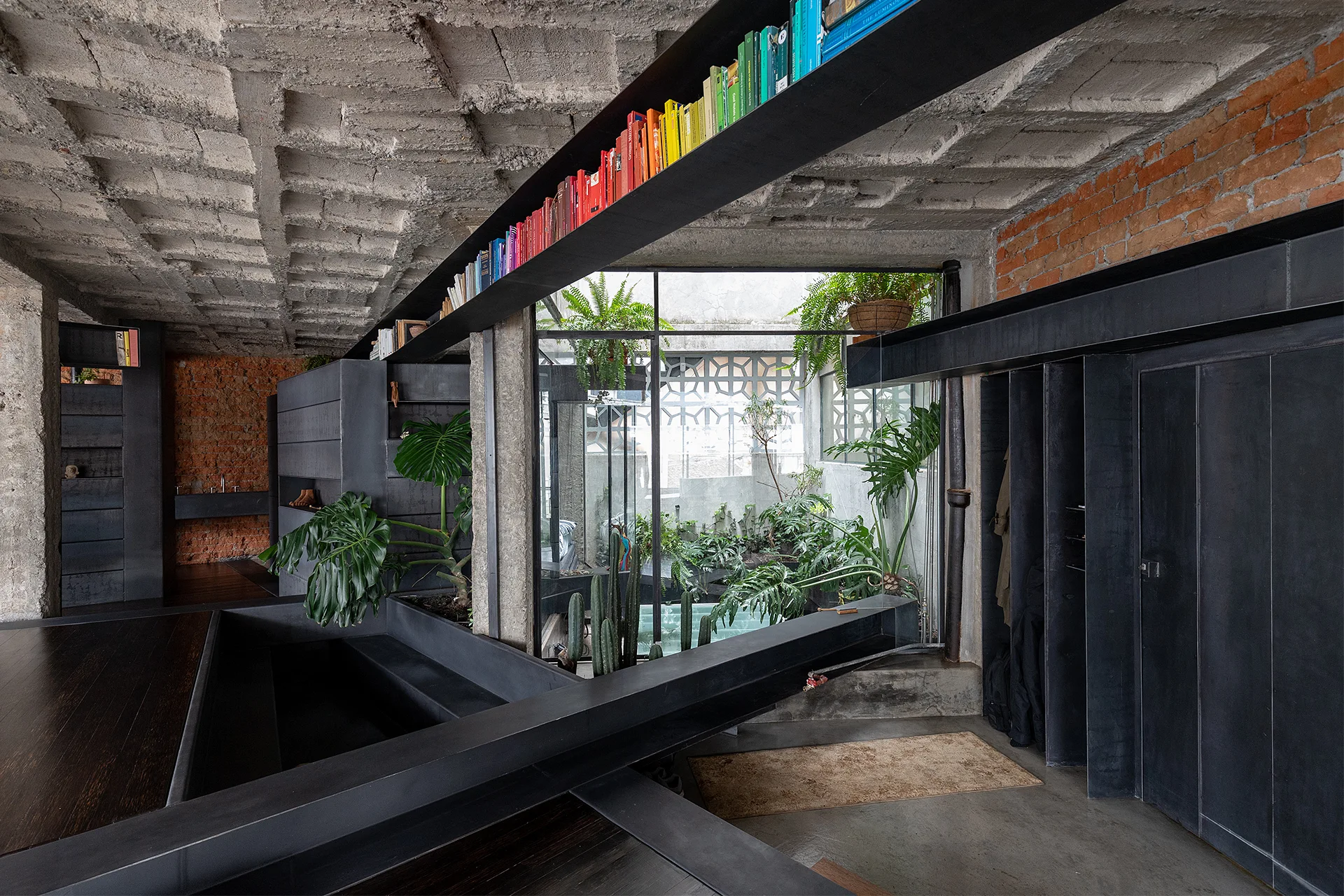 An industrial interior featuring a coffered concrete ceiling, black steel beams with books, and an enclosed courtyard with green plants.