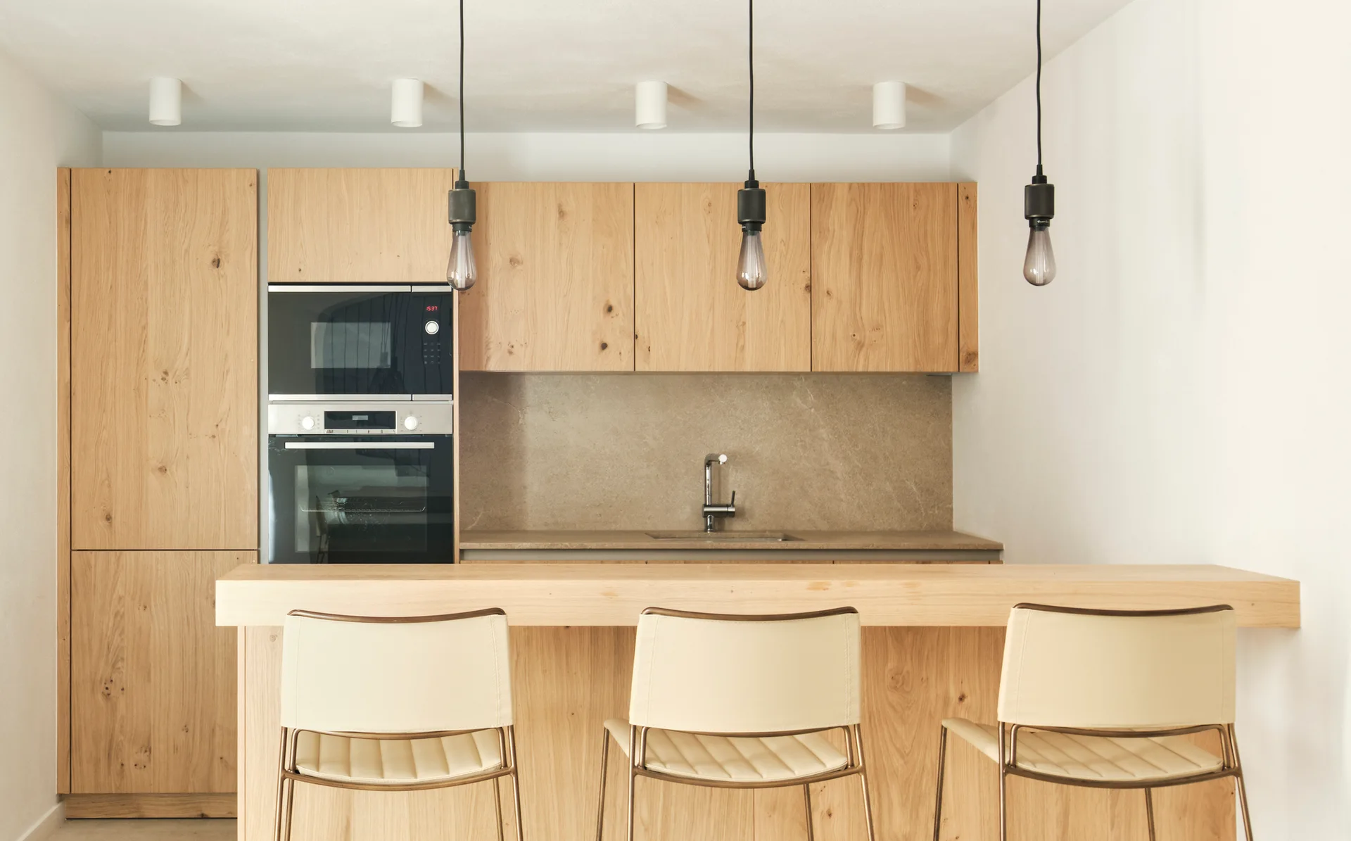 Three beige bar stools sit along a thick wood island in a minimalist kitchen featuring light oak cabinets, built-in ovens, and three pendant lights.