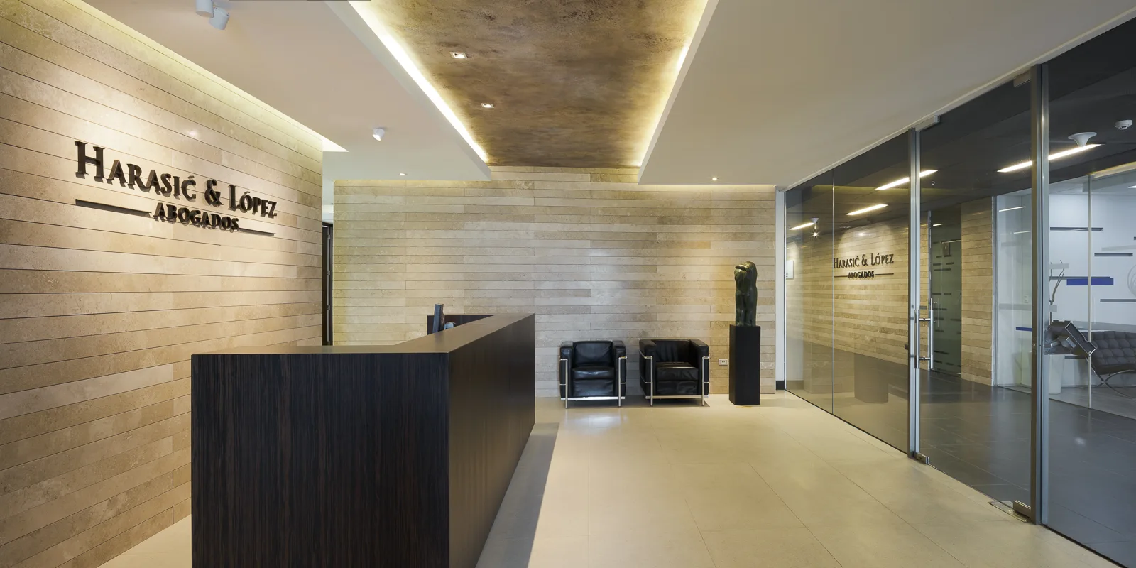 A modern office reception area featuring a dark wood desk, beige stone tiled walls, and two black leather armchairs near a glass partition.