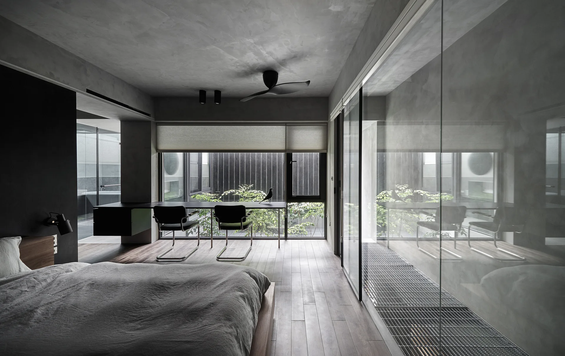 A minimalist bedroom with concrete walls, a wood floor, a platform bed, and a long black desk facing a window with a green courtyard view.