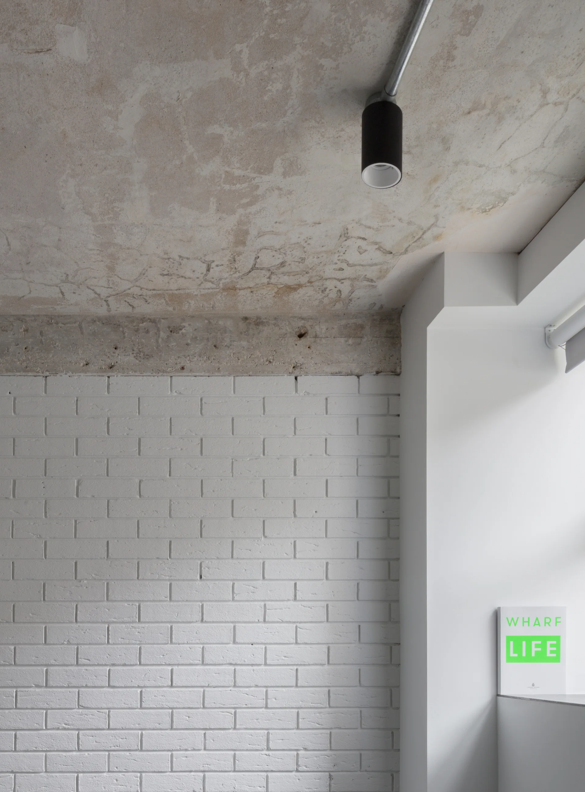 An industrial interior detail showcasing a white painted brick wall directly beneath a raw exposed concrete ceiling with a black spotlight.
