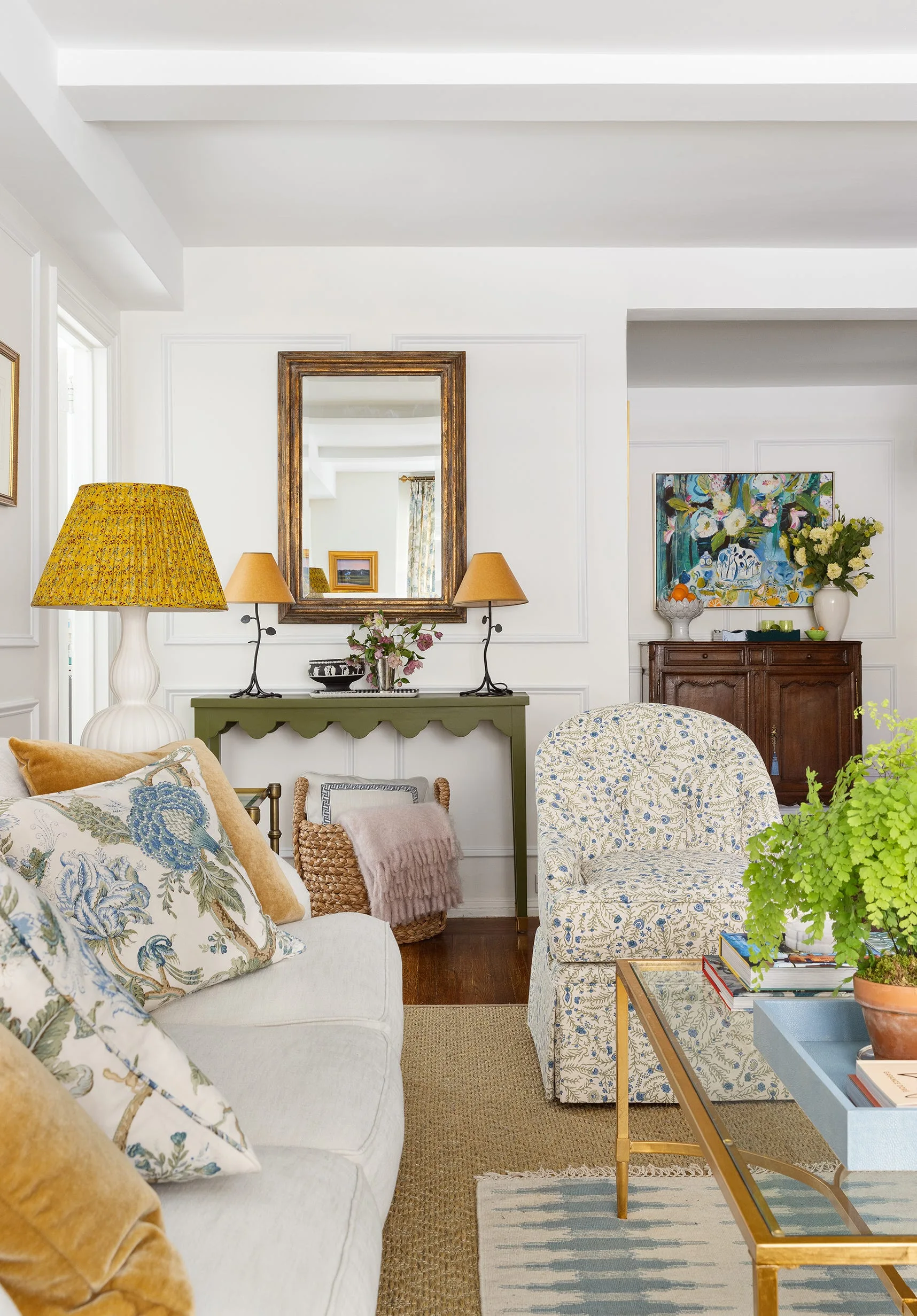 A bright living room features a white sofa, a blue floral armchair, and a scalloped green console table beneath a gold-framed mirror.