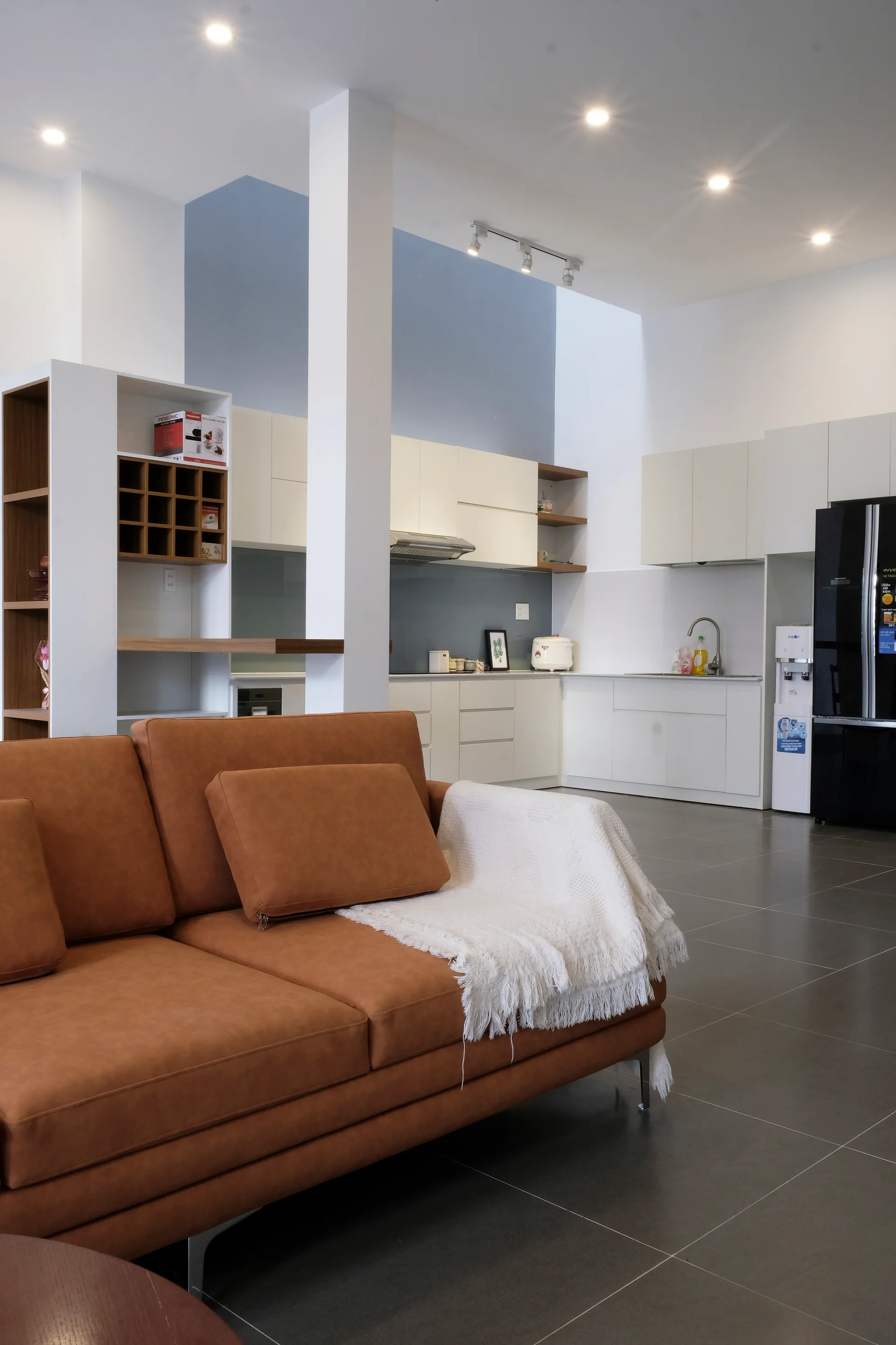 A tan leather sofa with a white throw blanket sits in the foreground of a modern open-plan kitchen featuring white cabinets and grey tiled floors.