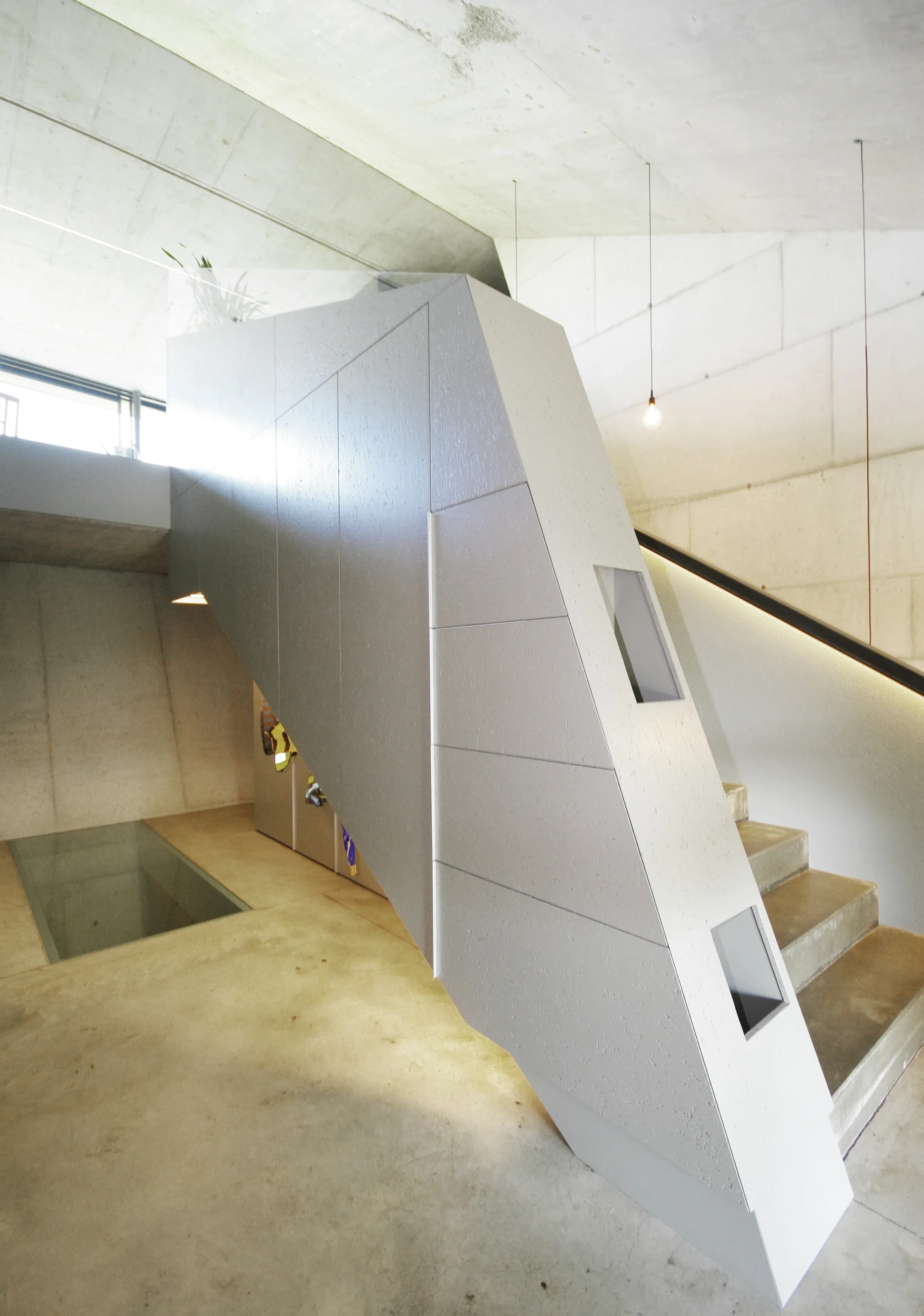 A modern concrete staircase featuring an angular, geometric gray enclosure, concrete block walls, and a glass panel inserted into the floor.
