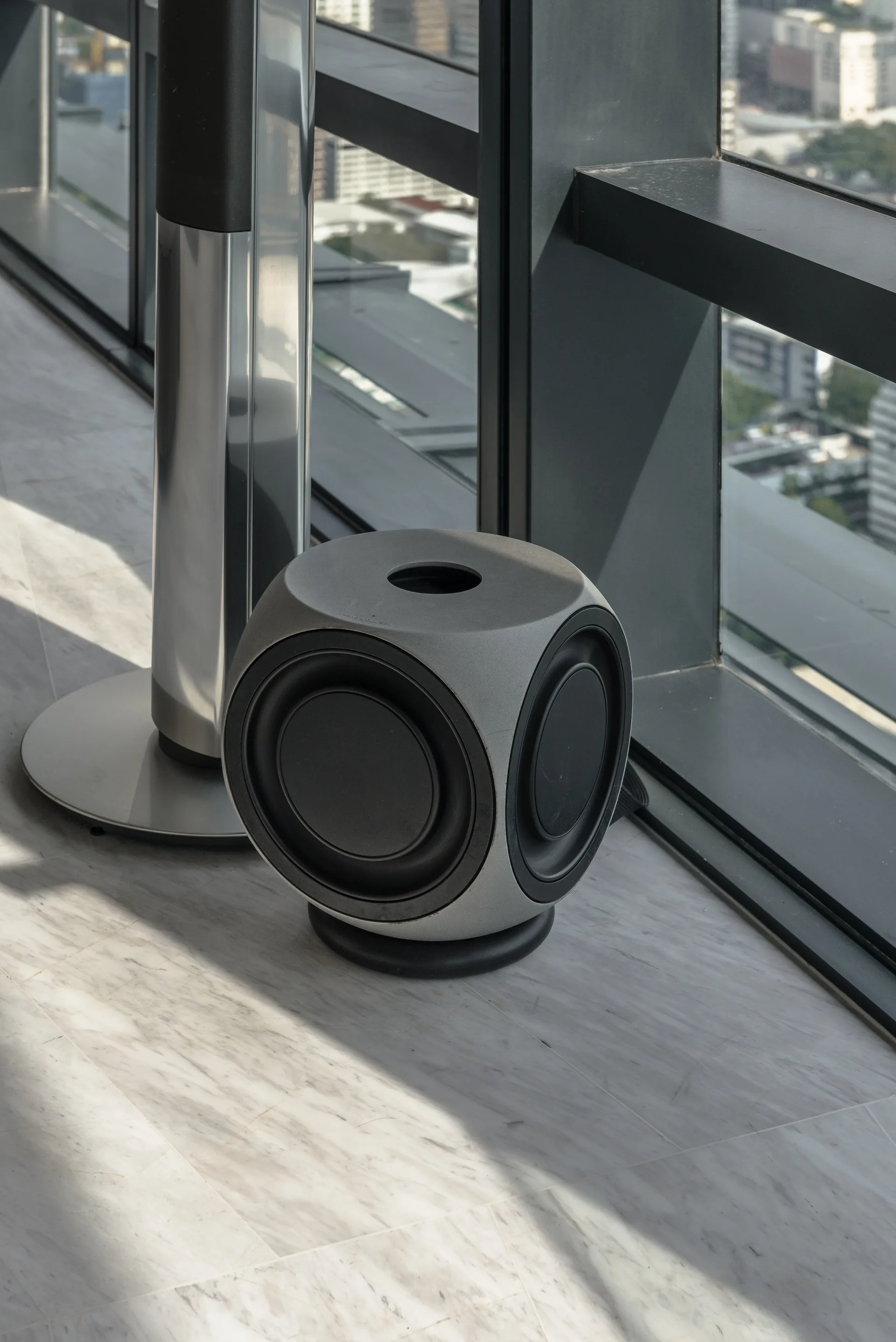 A grey cube-shaped subwoofer and a silver speaker column sit on a veined white marble floor near a dark window frame.
