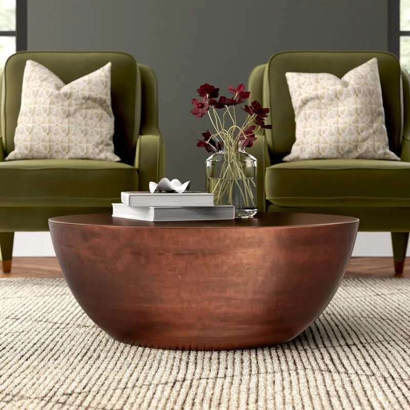 A round copper bowl coffee table sits on a striped neutral rug between two olive green armchairs with patterned pillows.
