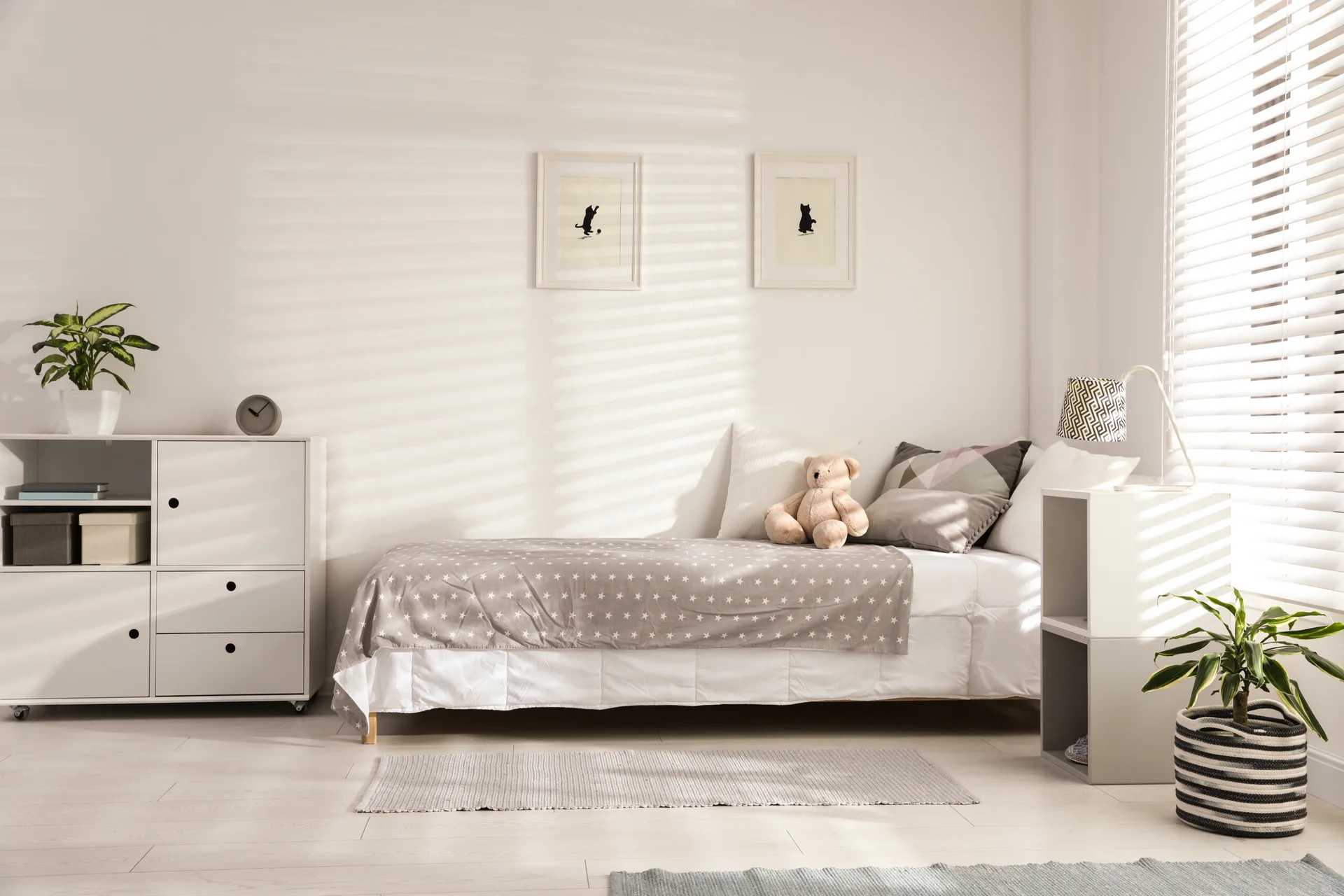 A kids bedroom features a single bed with star-patterned bedding, white modular cabinets, and natural light filtering through window blinds.