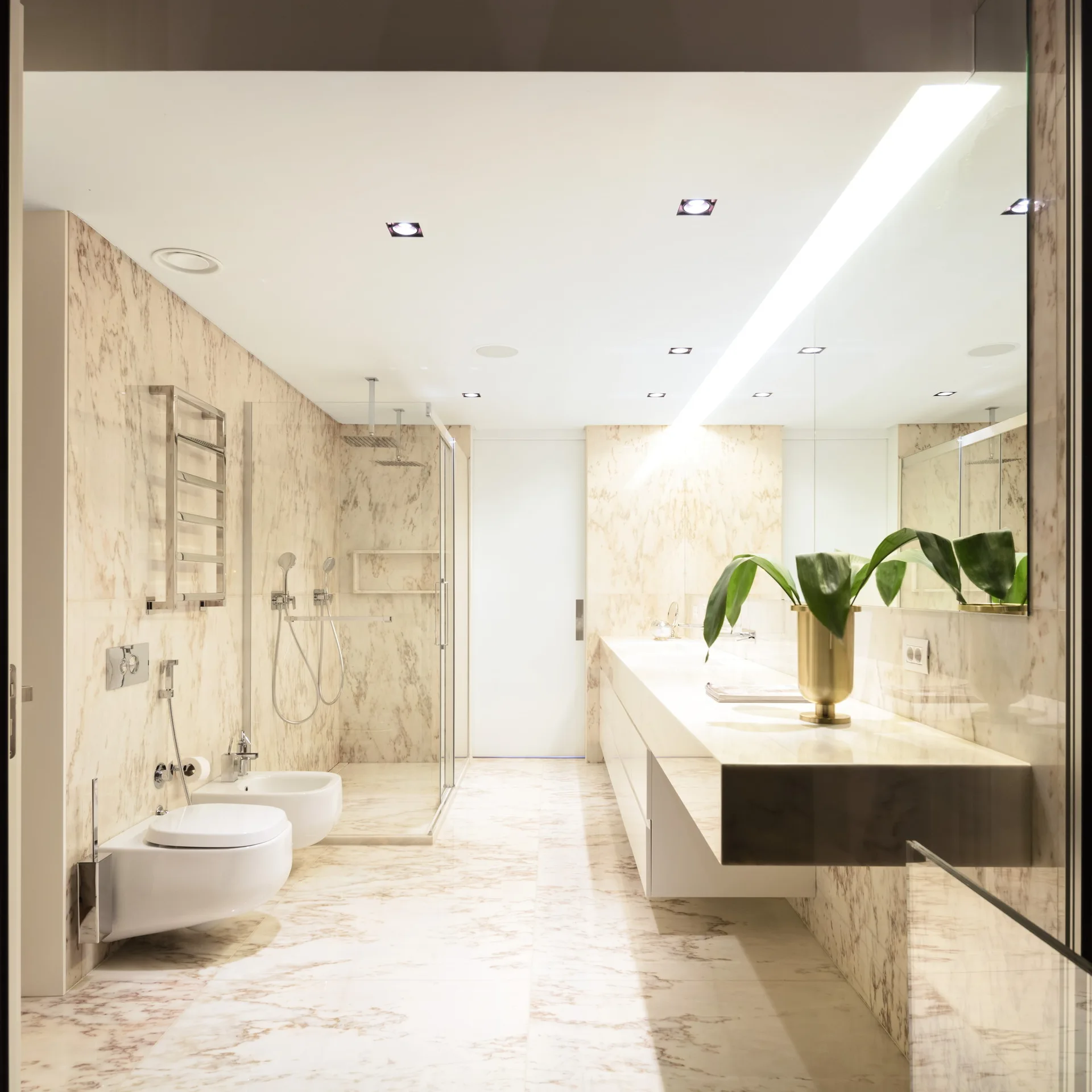 A modern bathroom features beige veined marble walls and floors, a long white floating vanity, and wall-mounted toilets next to a glass shower.