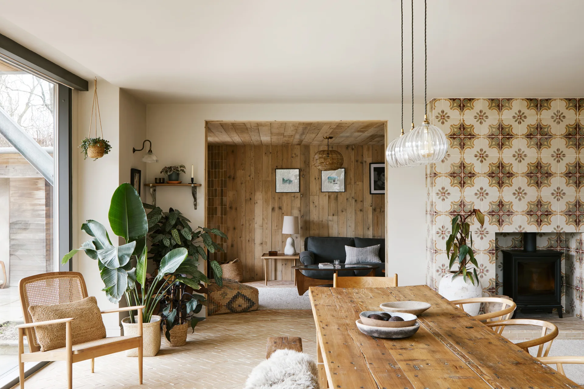 An open-plan dining area features a long wooden table, glass pendant lights, a patterned tile fireplace, and a wood-paneled alcove in the background.