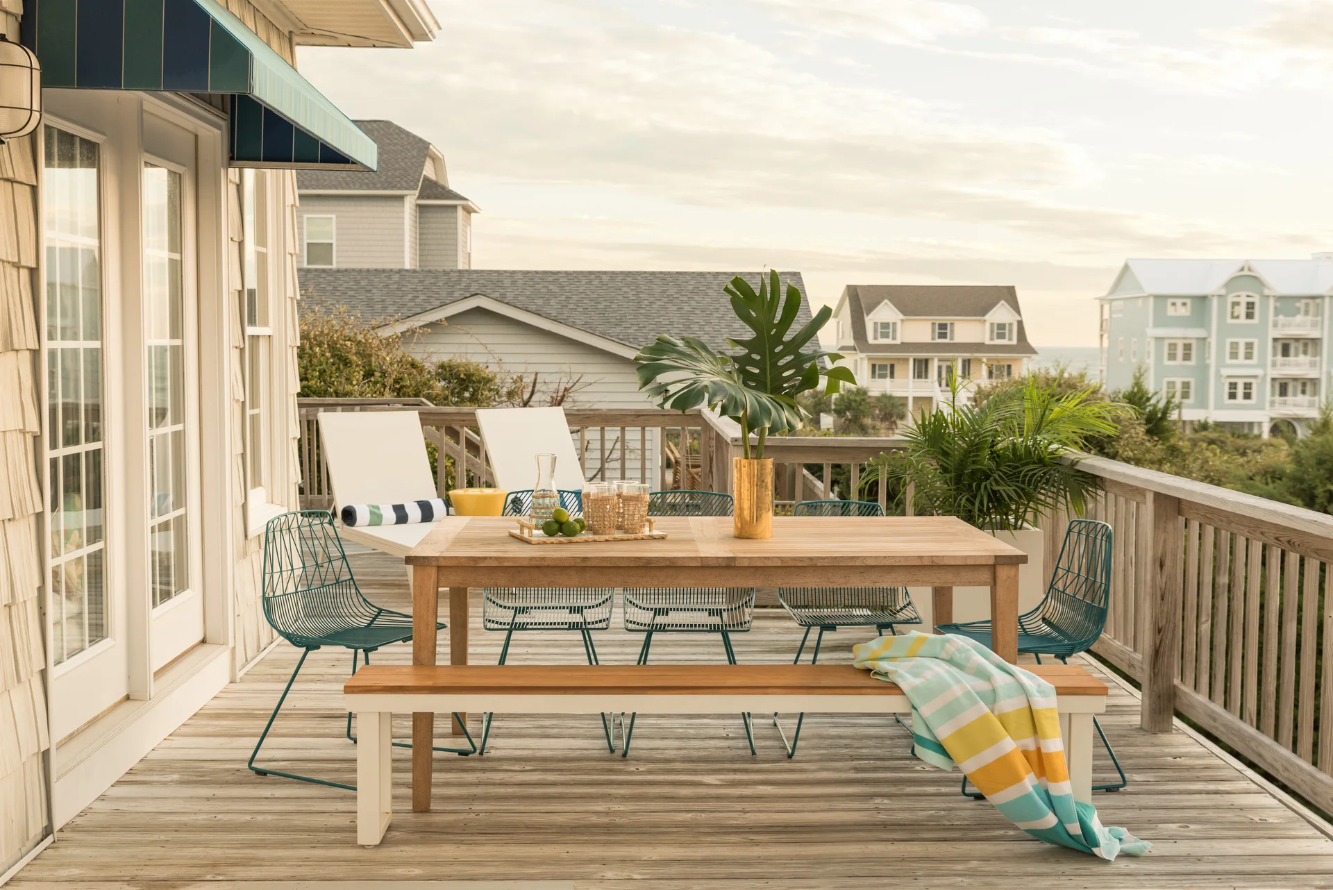 An outdoor wooden deck features a wood dining table surrounded by teal wire-frame chairs and a wooden bench, with coastal houses in the background.
