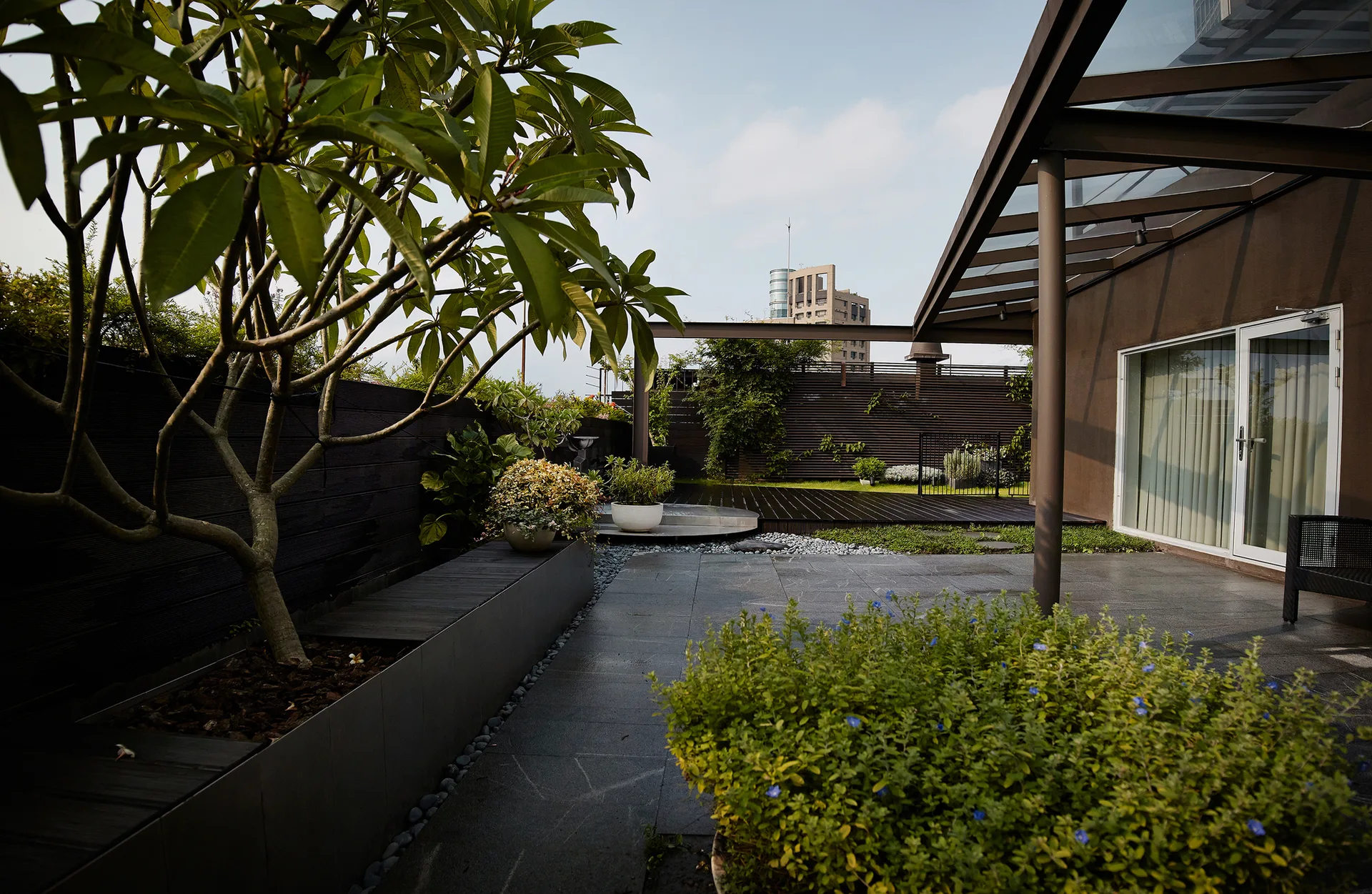 A modern outdoor terrace featuring dark stone tiled flooring, a glass-covered patio canopy, and a dark horizontal slatted privacy fence.