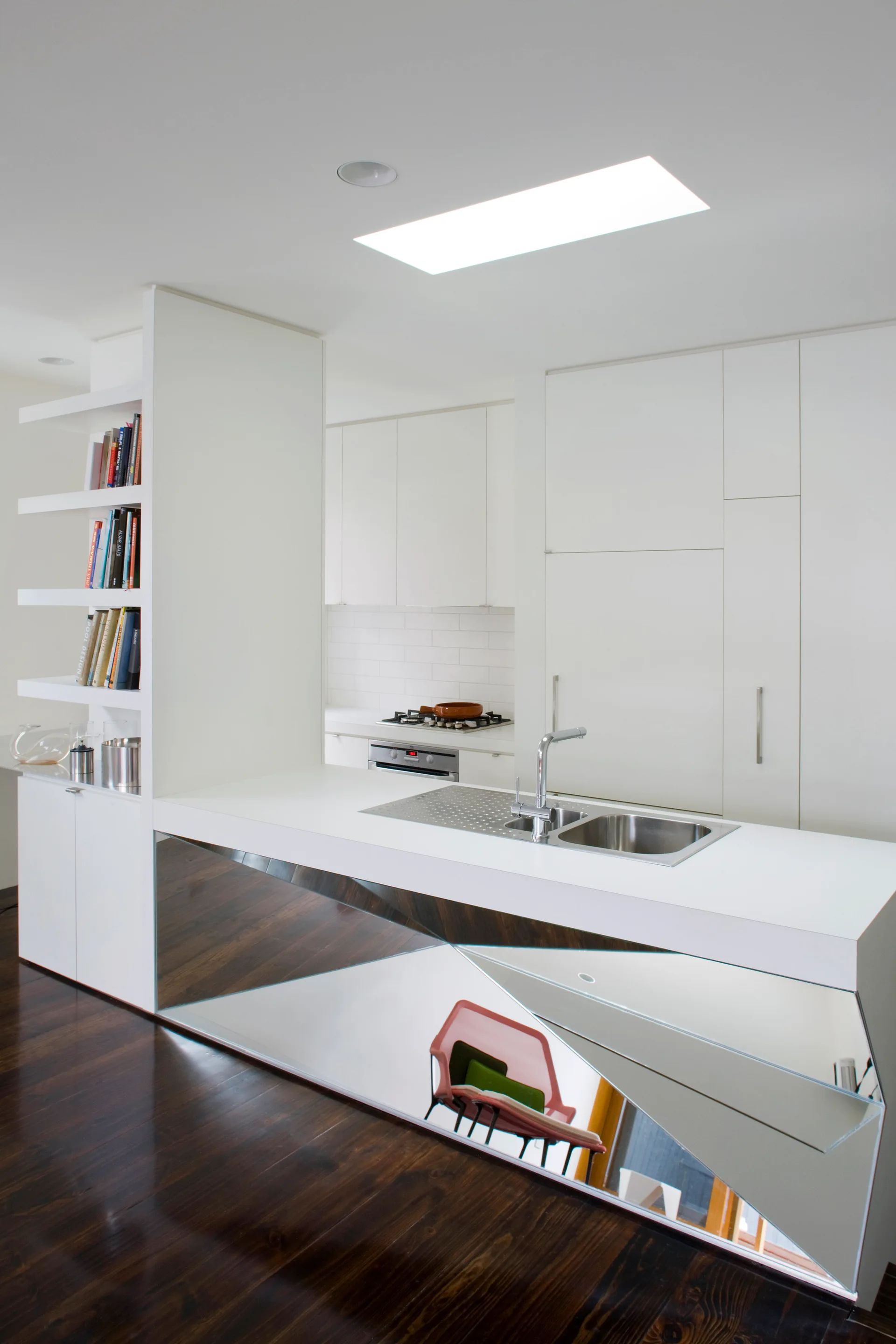 A minimalist white kitchen featuring an island with a faceted mirrored front, stainless steel sink, open shelves, and dark wood flooring.