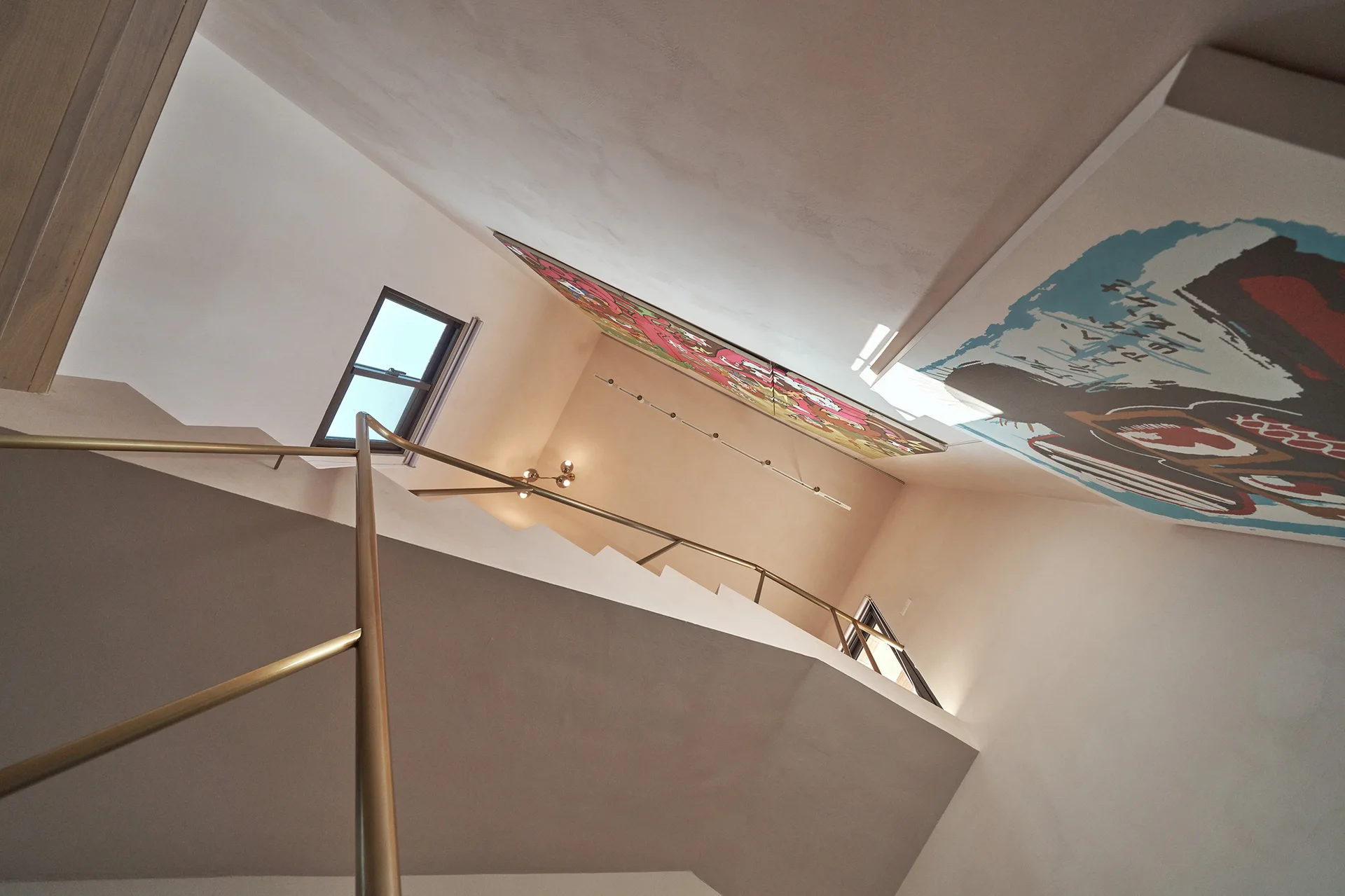 An upward-looking view of a modern stairwell with matte beige walls, a gold metal handrail, and colorful framed art hanging near the ceiling.