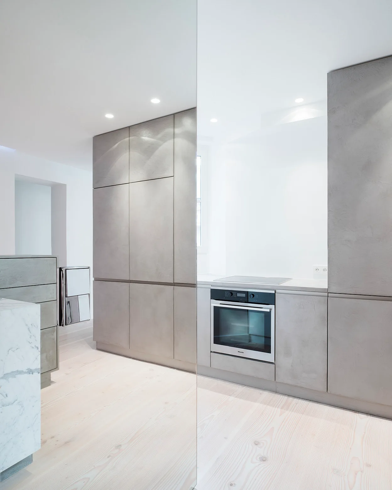 A minimalist kitchen features textured grey matte cabinetry with a built-in oven, pale wood plank flooring, and the corner of a white marble island.