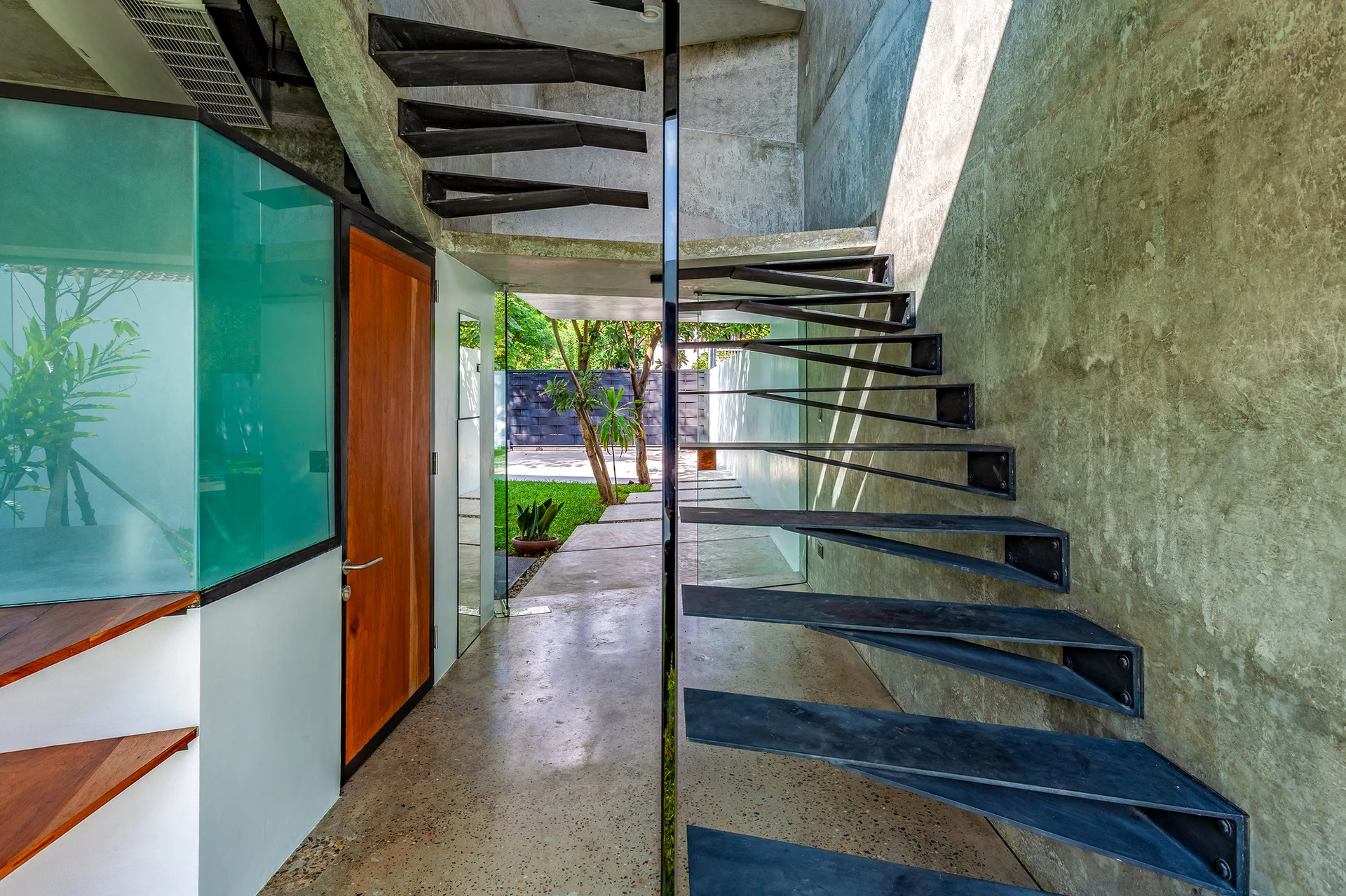 A floating black metal staircase rises along a textured raw concrete wall in a minimalist entryway leading to an outdoor garden.
