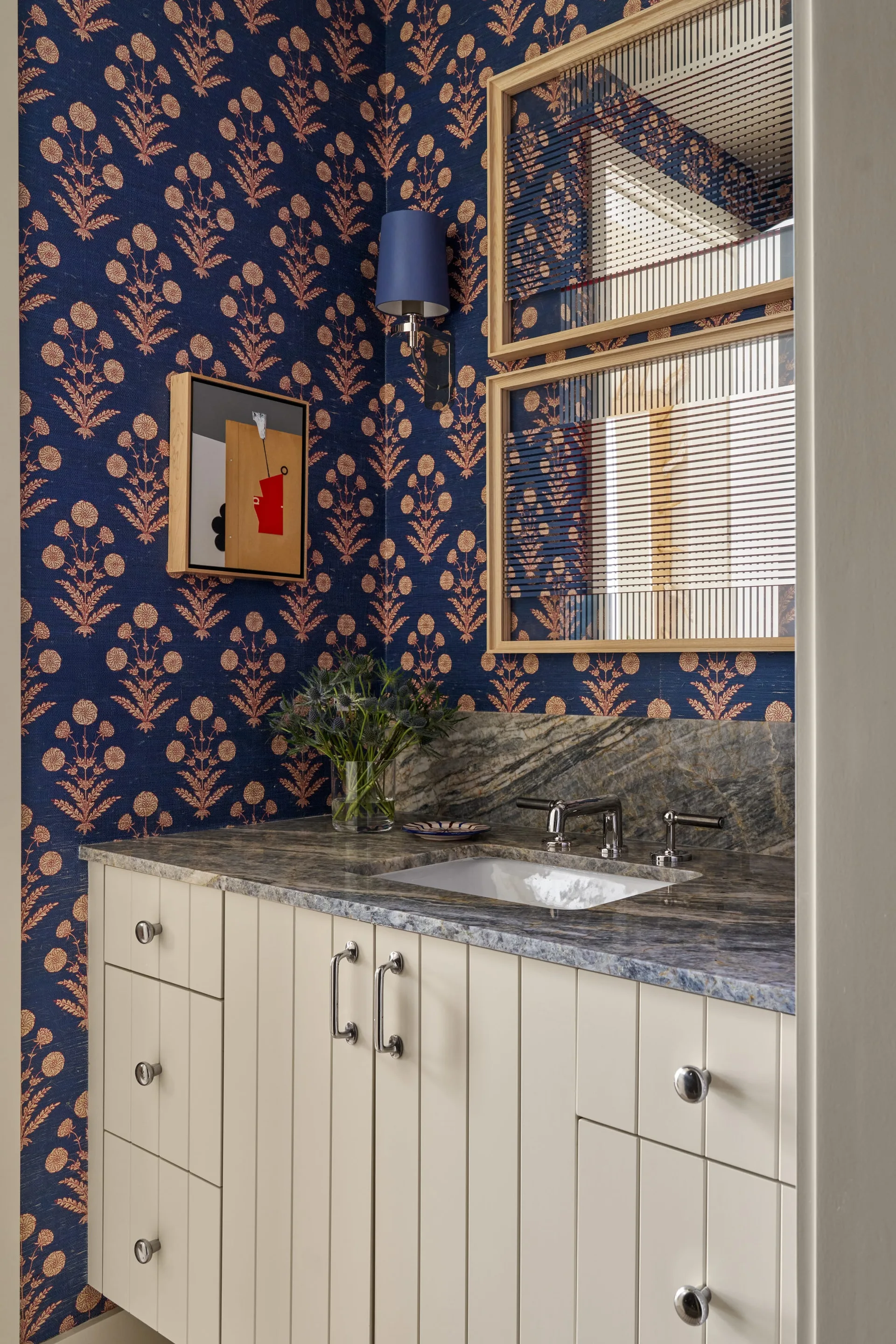 A powder room features a cream vertical-planked vanity with a veined stone countertop, set against navy blue floral wallpaper.