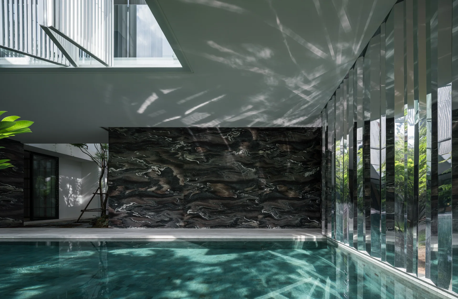 A modern swimming pool with a veined dark marble feature wall and vertical reflective panels under a white ceiling with a skylight.