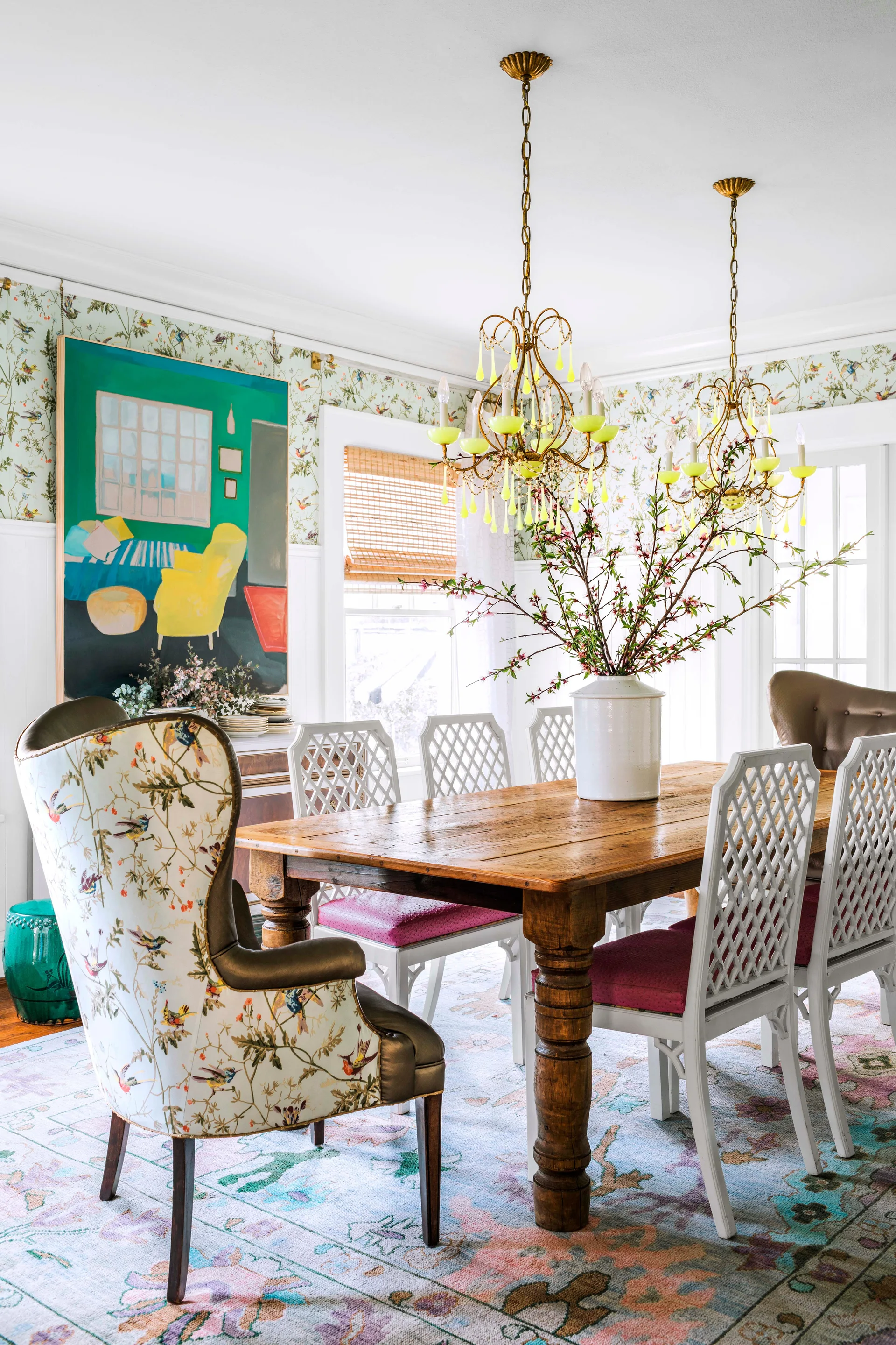 An eclectic dining room features a rustic wood table surrounded by white lattice chairs, floral bird wallpaper, and two gold chandeliers.