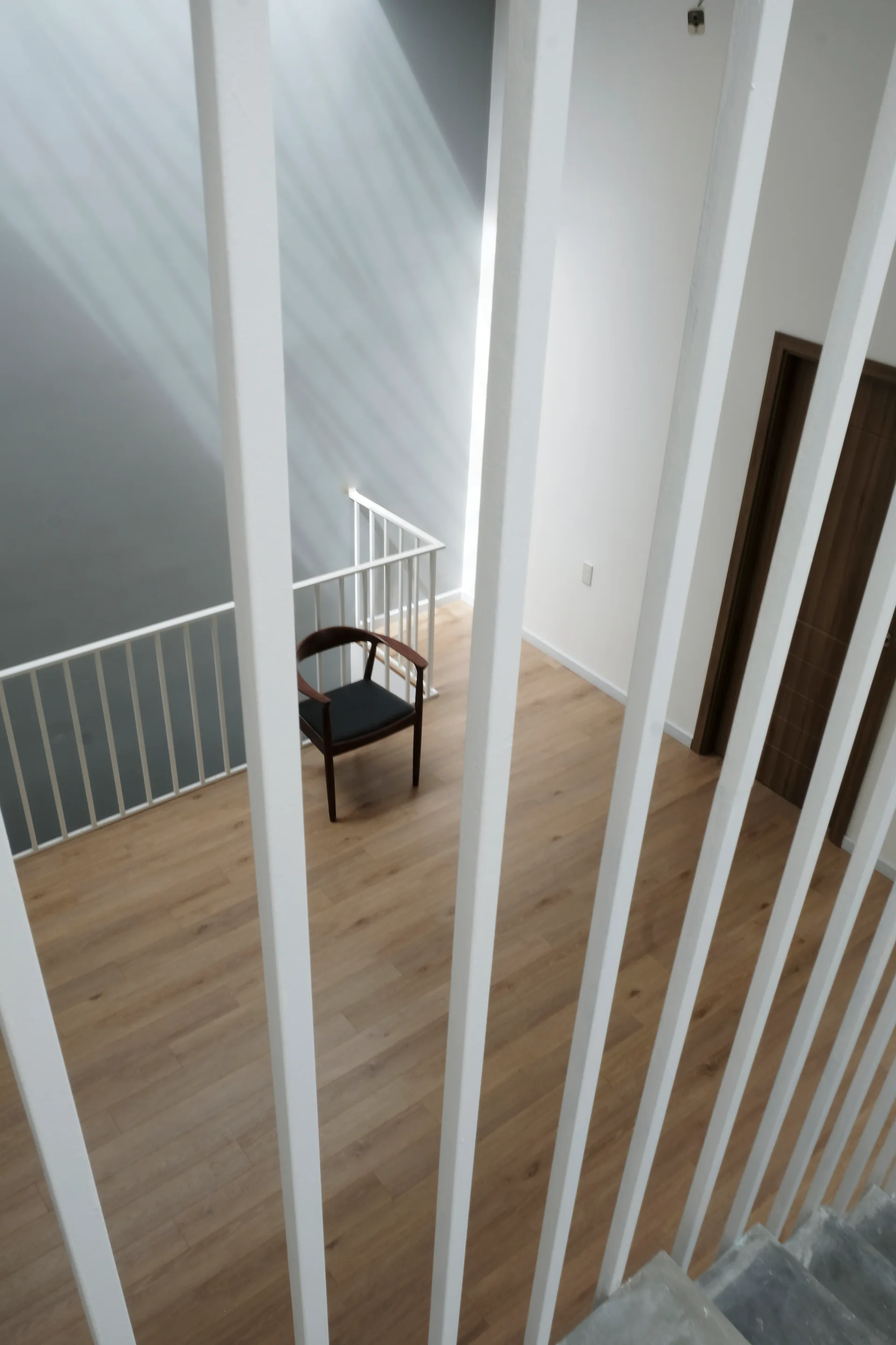 An overhead view through white vertical staircase railings onto a light wood landing with a single dark wooden armchair.