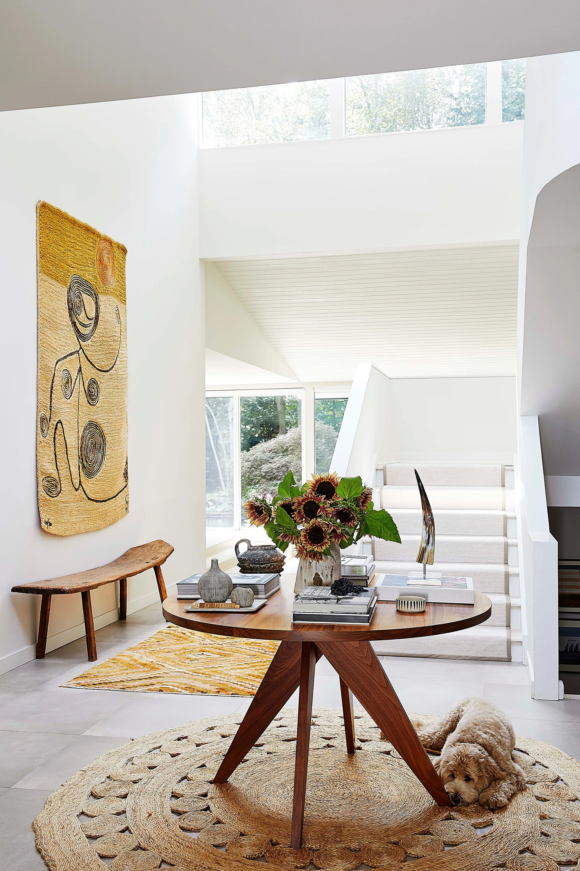 A bright white entryway features a central round wooden table on a circular woven rug, a wooden bench, a large tapestry, and stairs in the background.