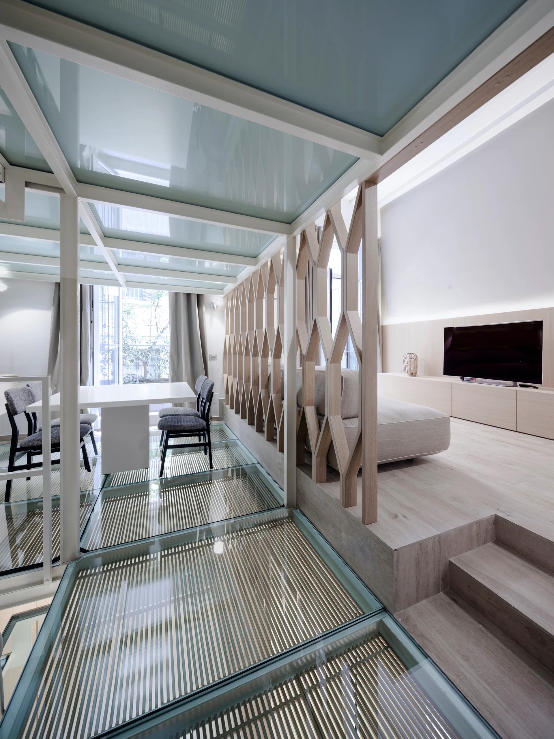 A modern split-level interior featuring a glass floor catwalk, a geometric light wood screen, and a raised living area with a beige sofa.