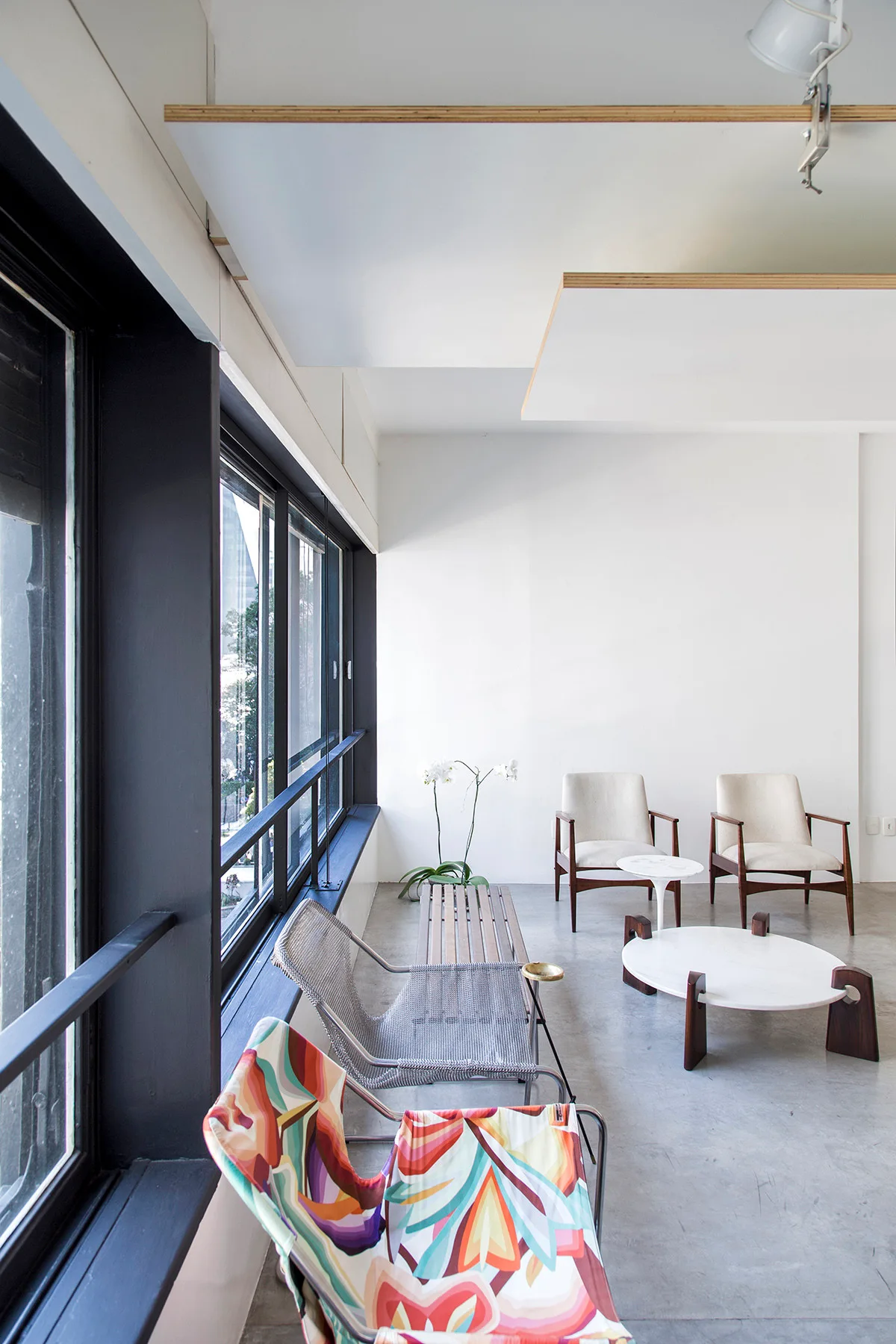 A minimalist modern room features mixed accent chairs, a slatted wood bench, and a round white coffee table beside large black-framed windows.