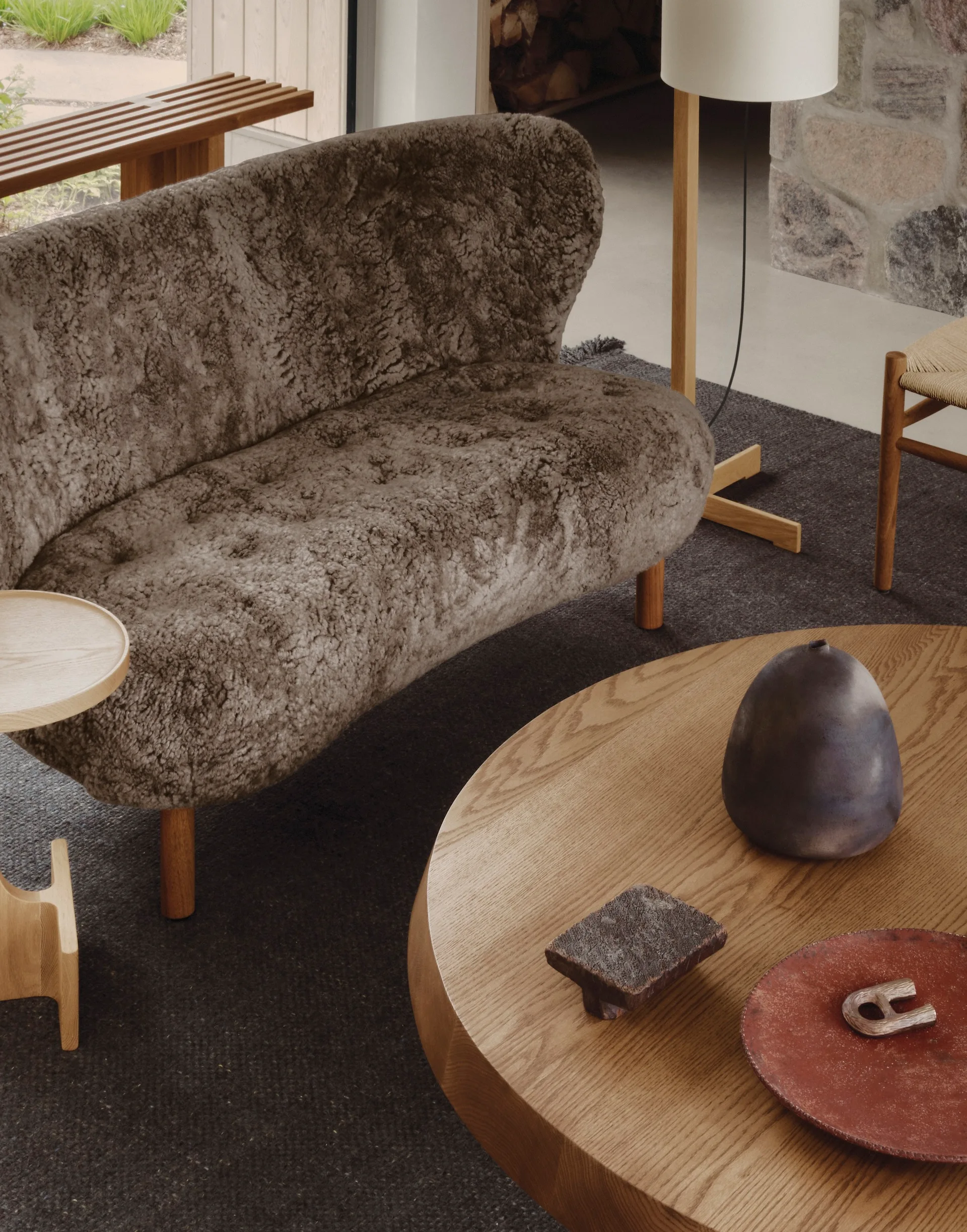 A textured shaggy brown sofa sits on a dark rug next to a round wooden coffee table styled with earthy ceramic vessels.
