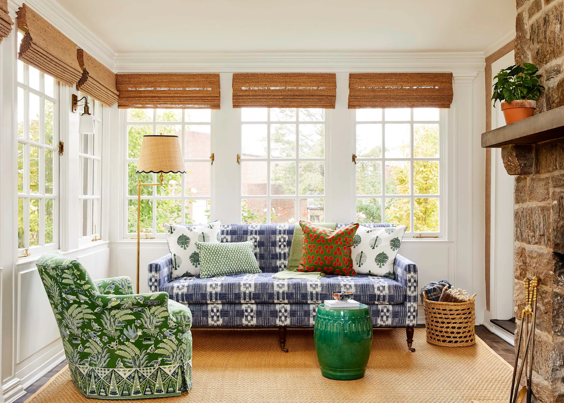 A bright living room features a blue patterned sofa, a green upholstered armchair, three windows with woven shades, and a rustic stone fireplace.