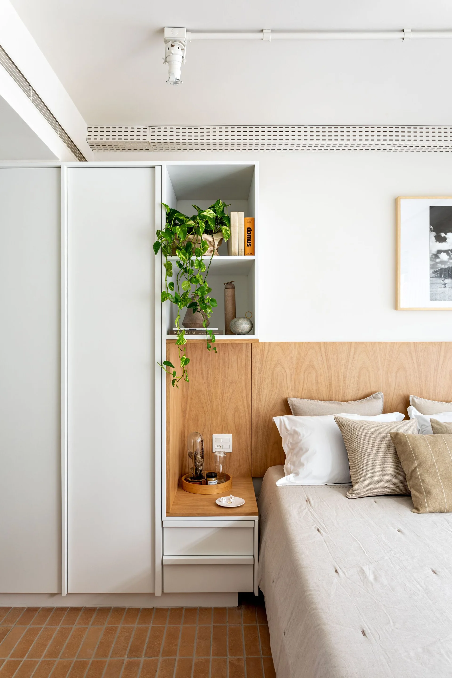 A bed with beige linen bedding sits next to a custom white wardrobe and integrated light wood headboard featuring a built-in display shelf.
