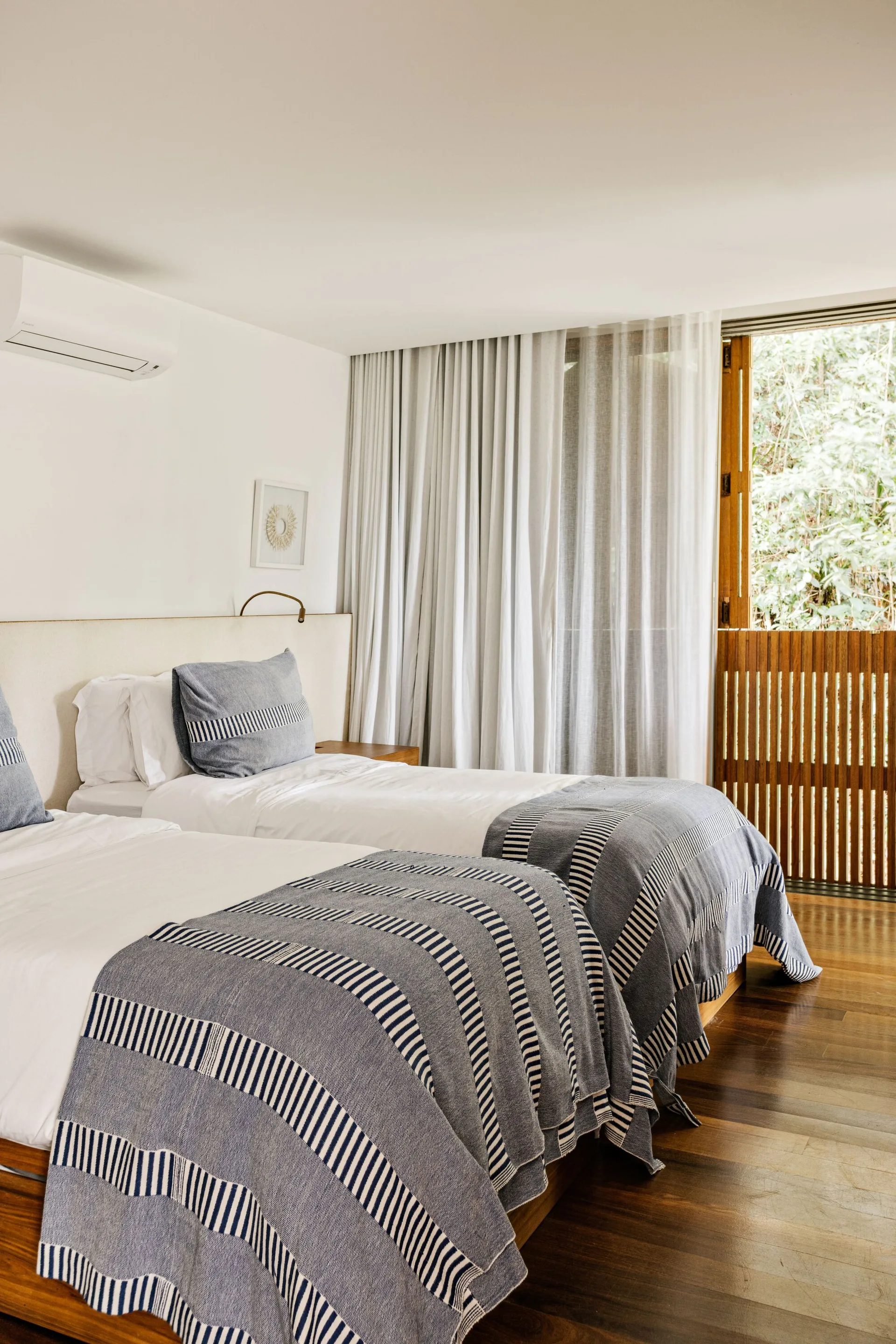 A bright bedroom with two beds featuring blue striped throws, a white headboard, white sheer curtains, and polished wood floors.