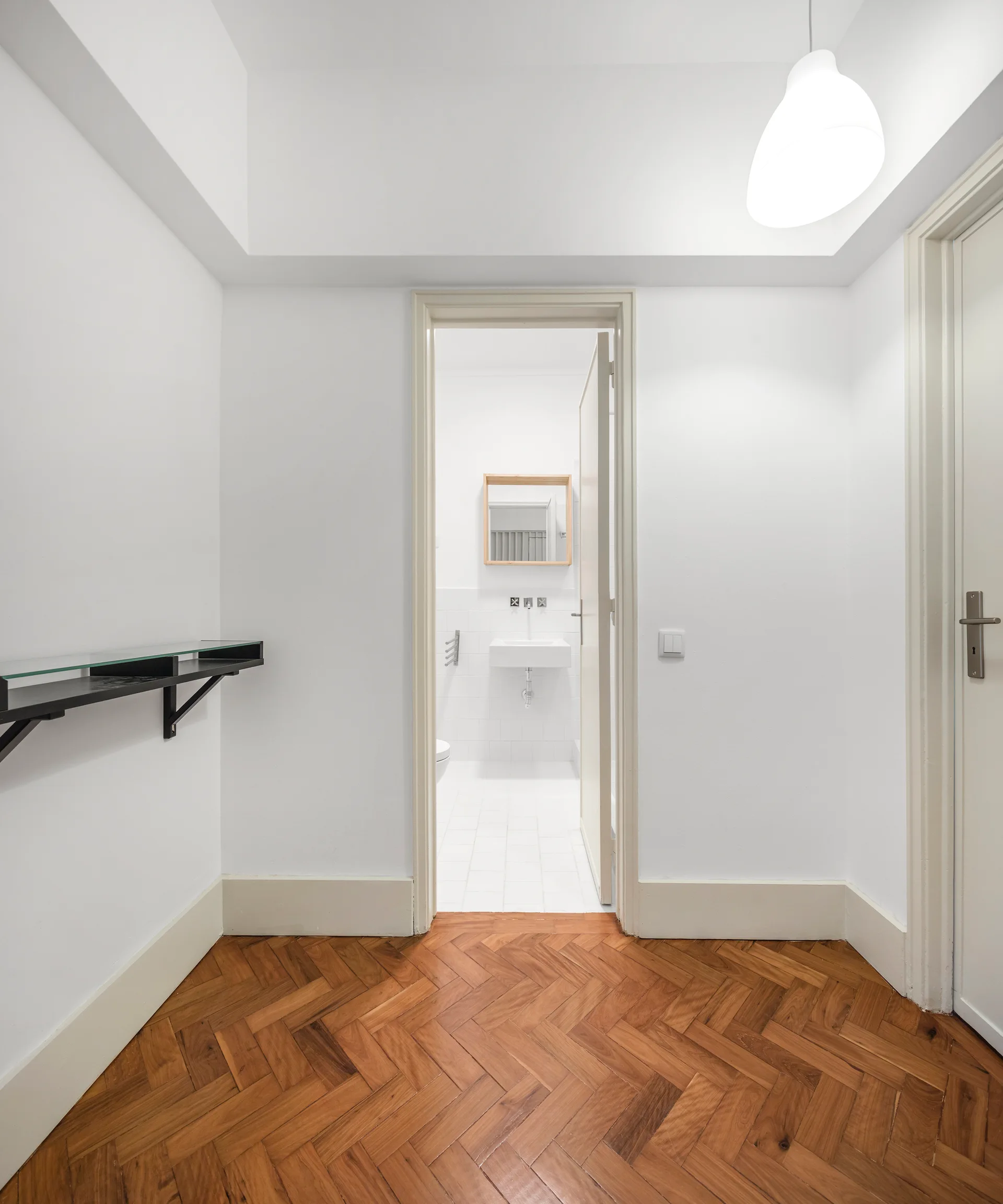 An open doorway connects a white hallway with herringbone wood flooring to a minimalist bathroom featuring a floating white sink and wood-framed mirror.