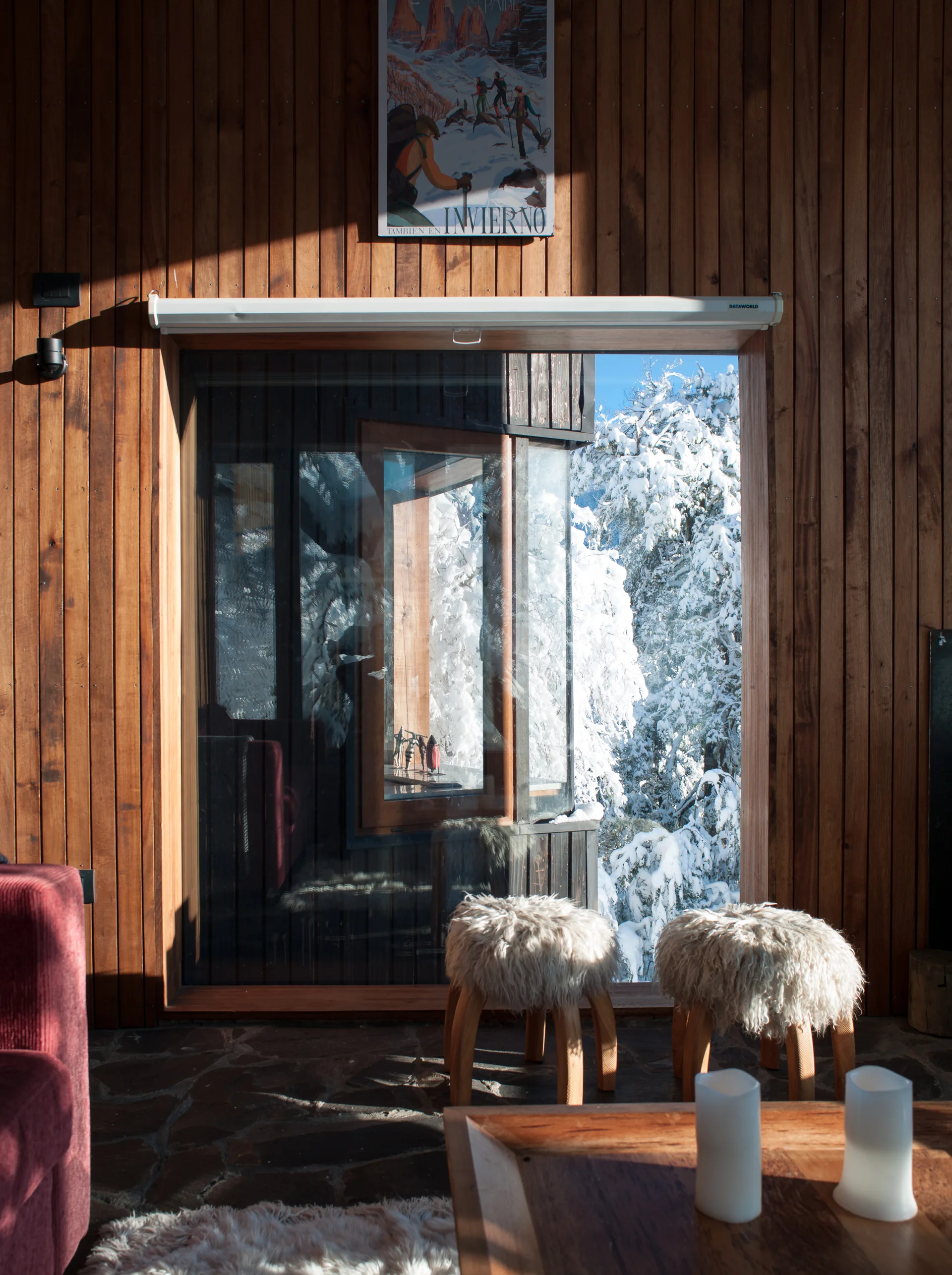 A rustic cabin living room with vertical wood paneling, a large window framing snowy trees, and two shaggy white stools on a flagstone floor.