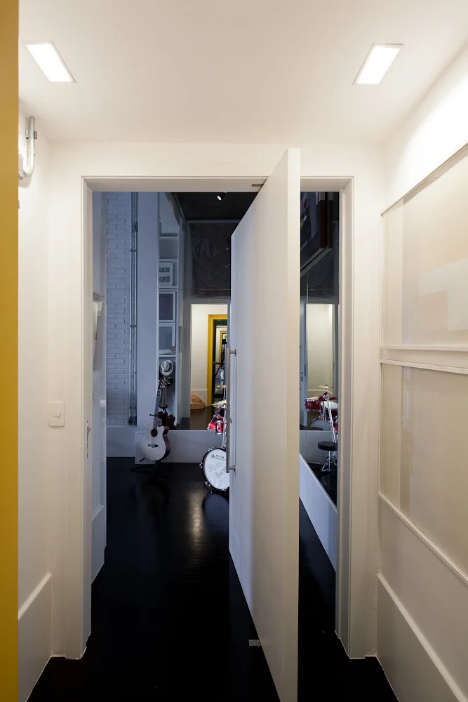 A large white pivot door stands partially open in a minimalist white hallway with a satin black floor and recessed ceiling lights.