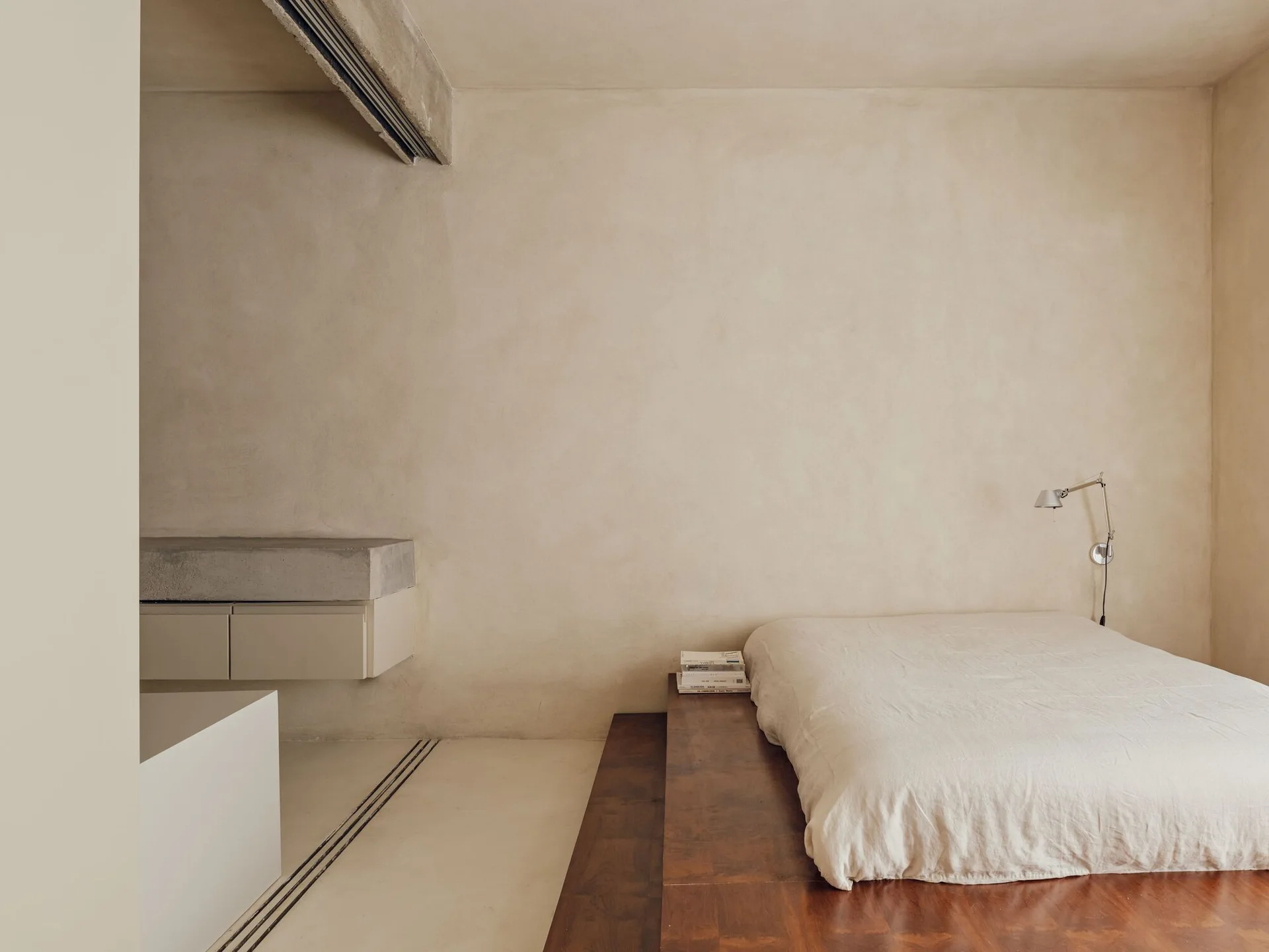 A minimalist bedroom featuring a low dark wood platform bed with cream linen bedding set against beige plaster walls.