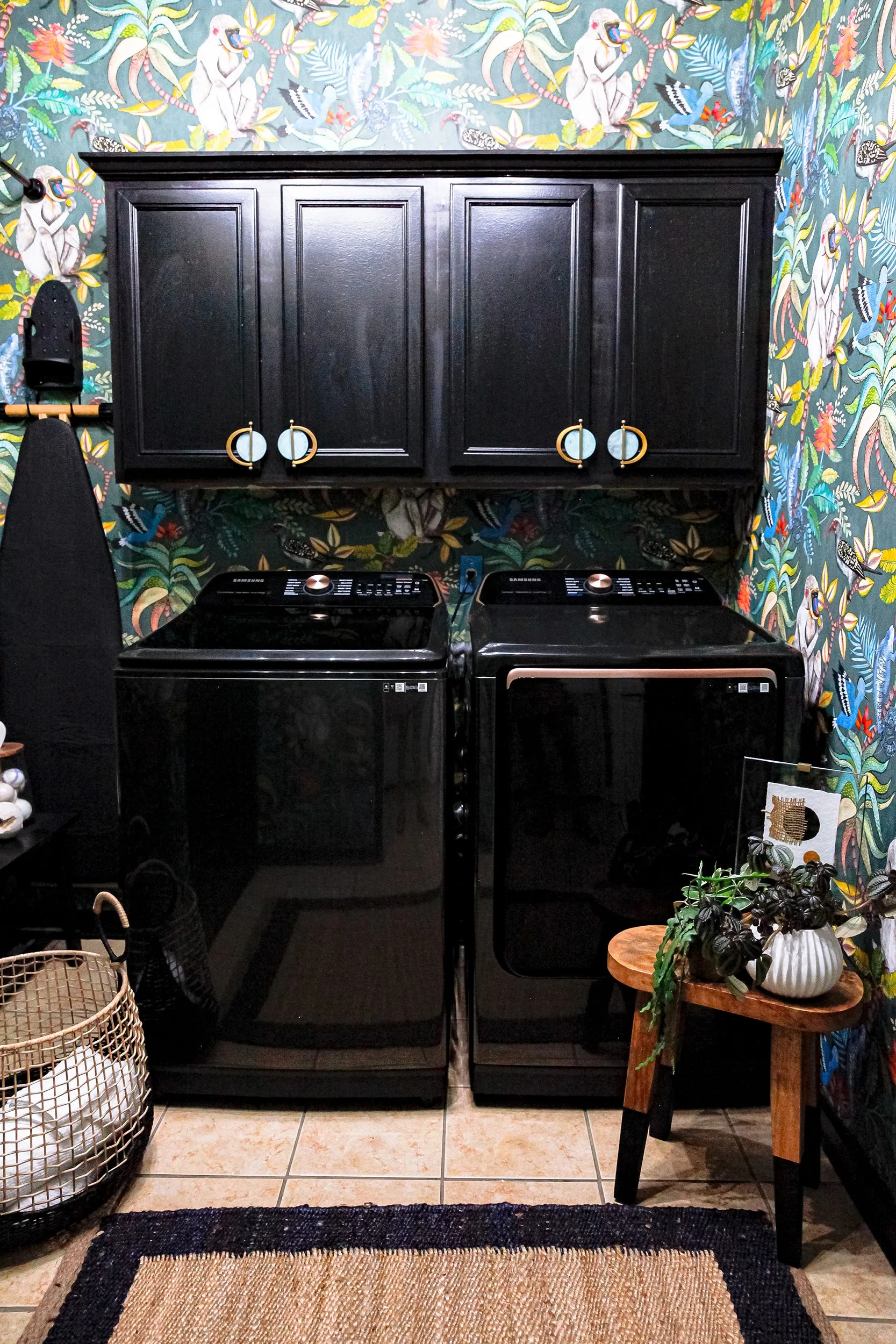 A utility room features black washing and drying machines under matching black cabinets set against a vibrant green jungle-patterned wallpaper.