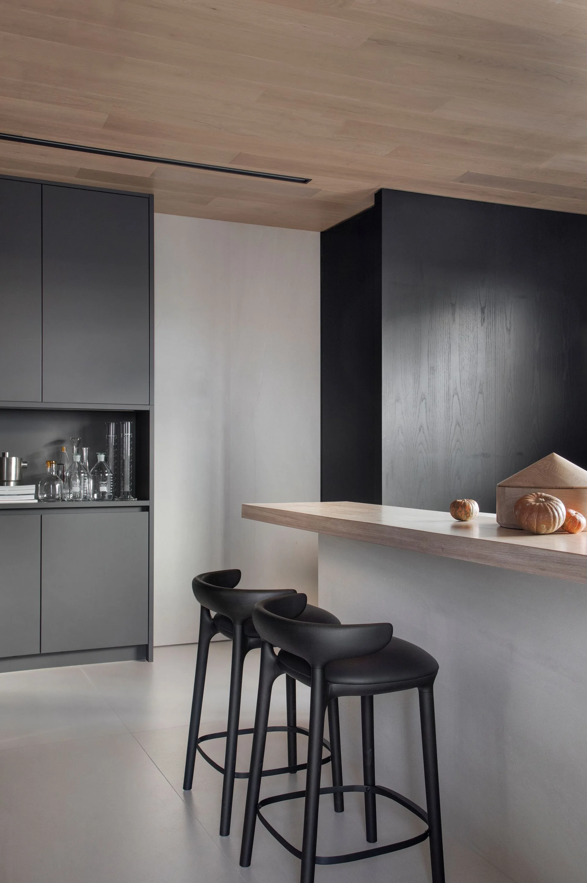 A minimalist kitchen features matte gray cabinetry, a concrete island with a wood countertop, black bar stools, and a wood plank ceiling.