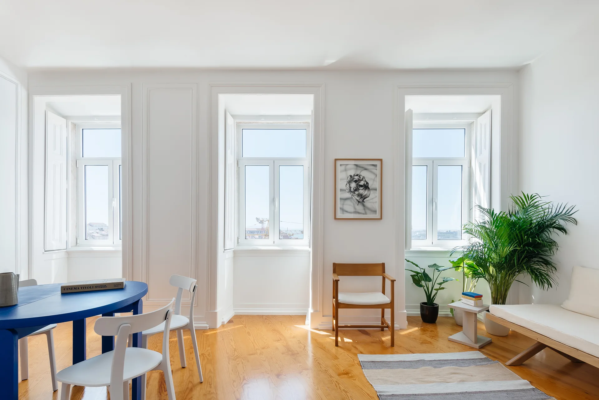 A bright dining room featuring a round cobalt blue table with white chairs, tall windows with interior white shutters, and warm wooden flooring.