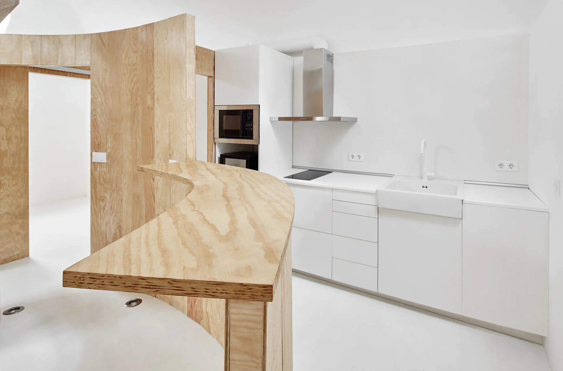 A minimalist white kitchen featuring a curved plywood breakfast bar, clean white cabinetry, built-in appliances, and a white sink.