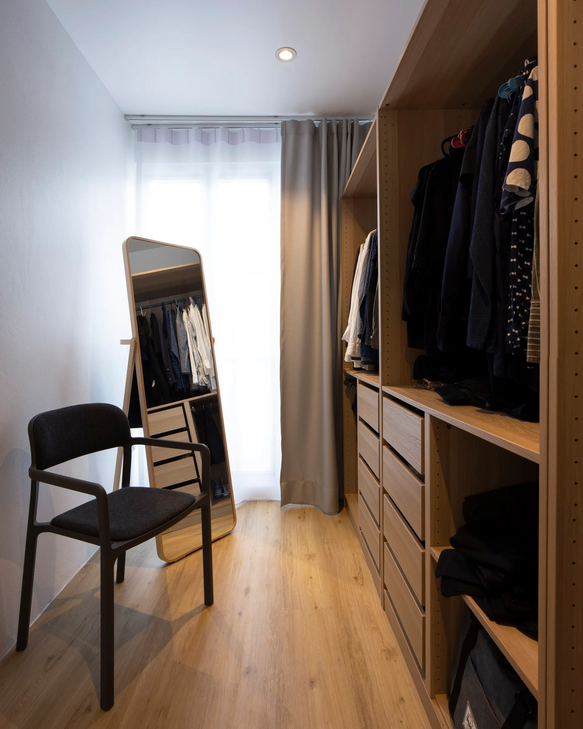 A minimalist walk-in closet featuring light wood built-in wardrobes, a floor-length standing mirror, a dark modern armchair, and a window.