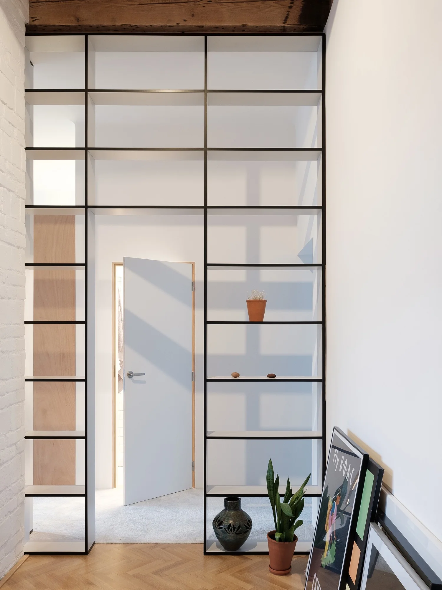 A black metal floor-to-ceiling shelving unit acts as a room partition in a hallway with herringbone wood floors and a white door.