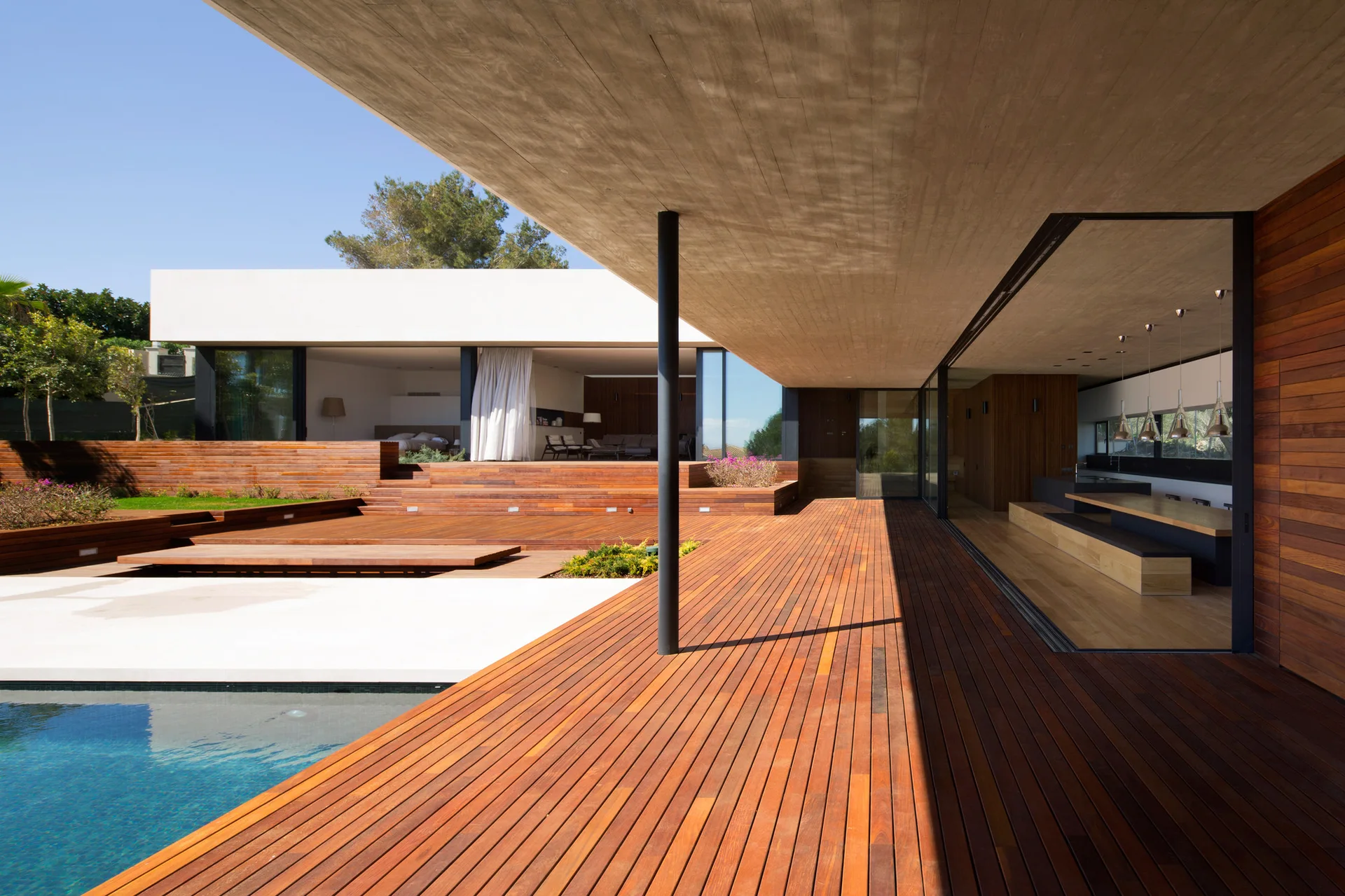 A modern concrete and wood patio features an expansive wooden deck alongside a swimming pool beneath a large overhanging ceiling.