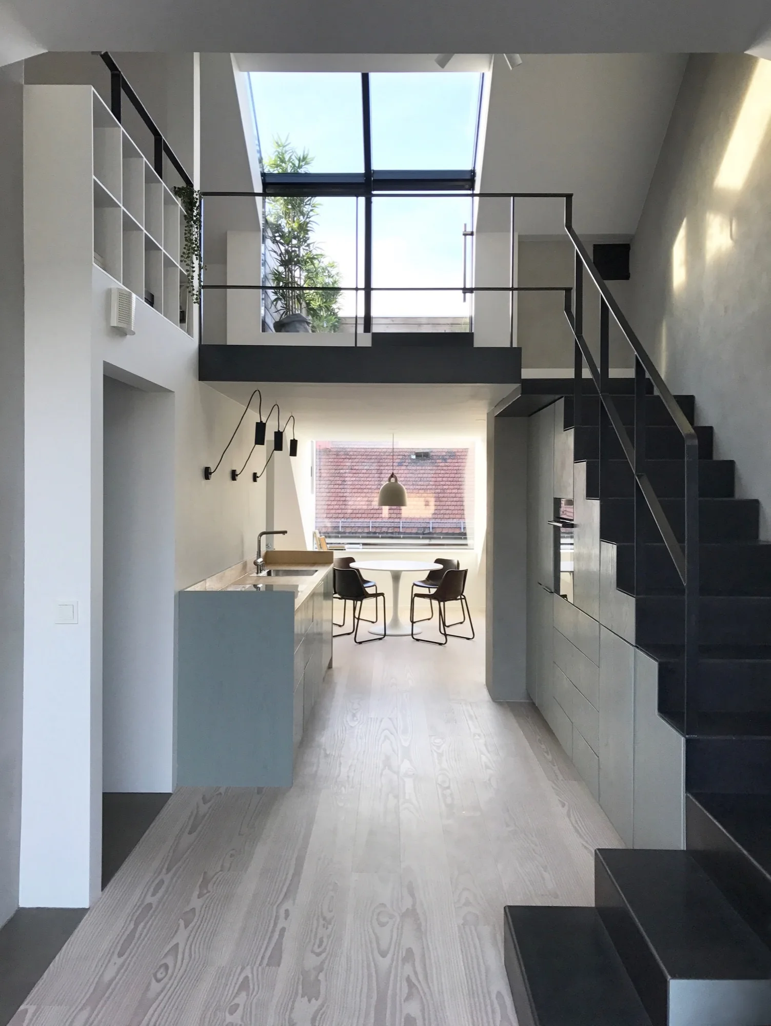 A modern loft kitchen featuring light wood floors, grey cabinetry, a black steel staircase, and a mezzanine level under a bright skylight.