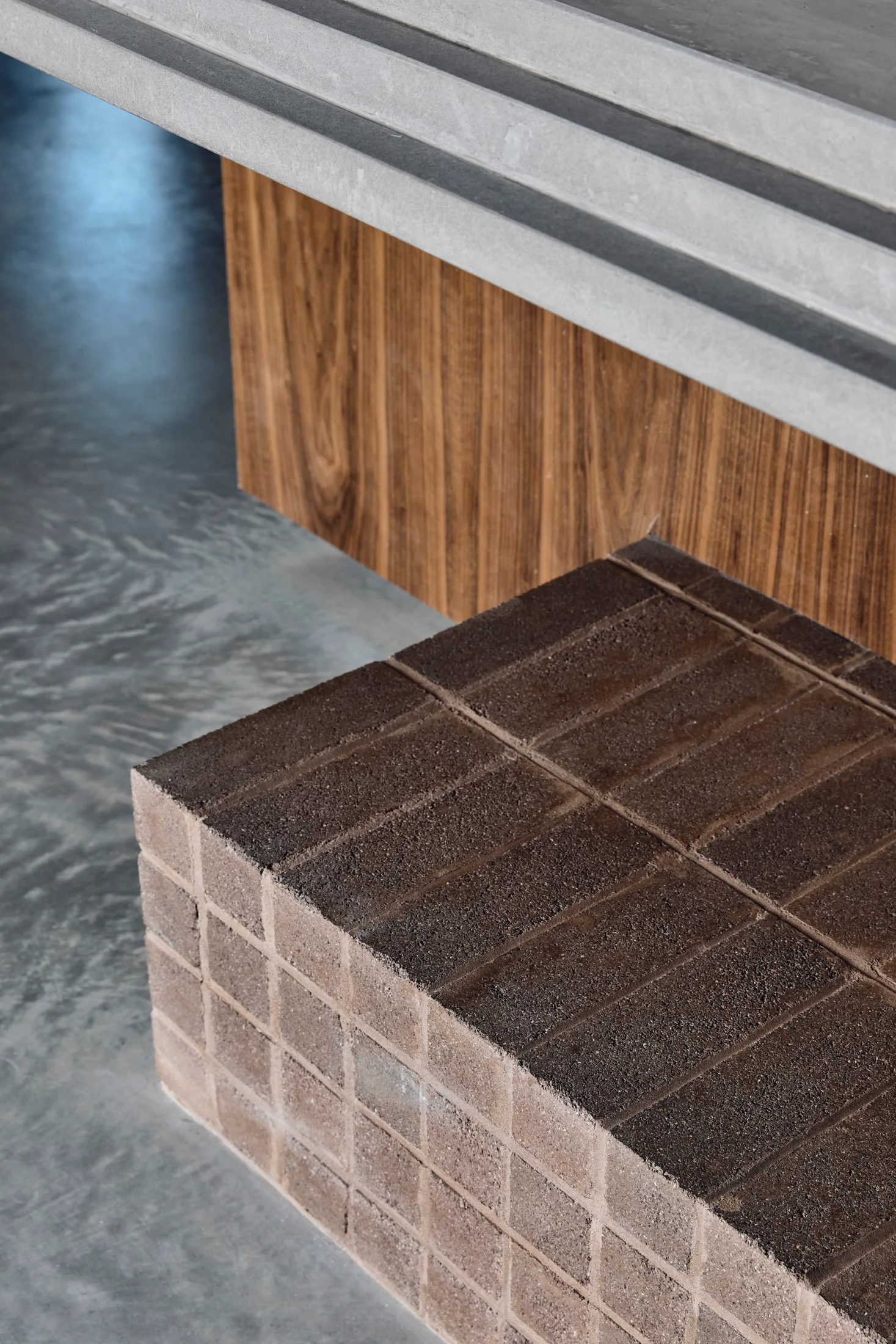 A close-up view of a textured brown stone block next to warm wood cabinetry underneath a layered grey concrete countertop.