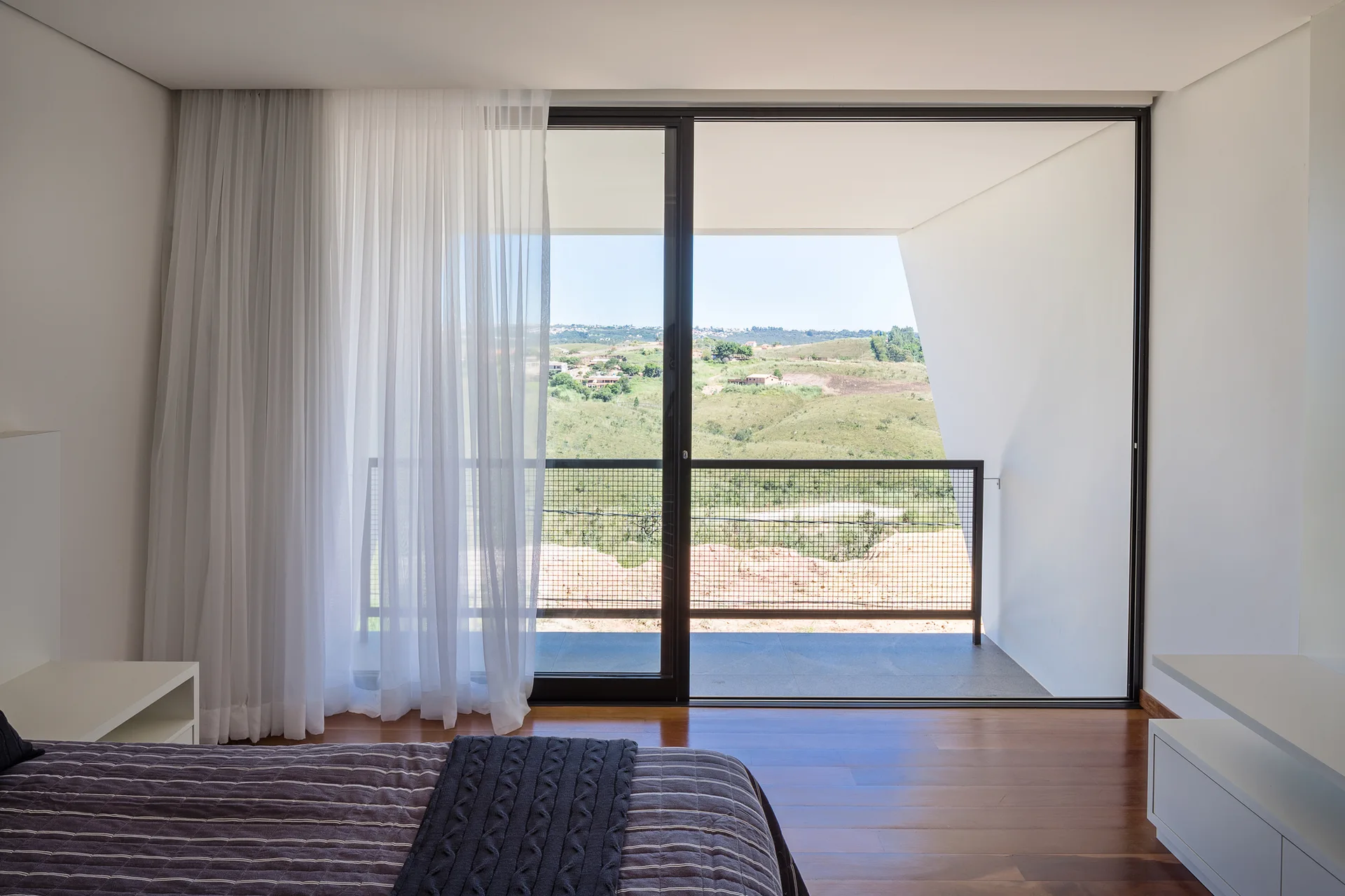 A minimalist bedroom features dark wood flooring, white walls, a bed with grey bedding, and large sliding glass doors leading to a balcony.