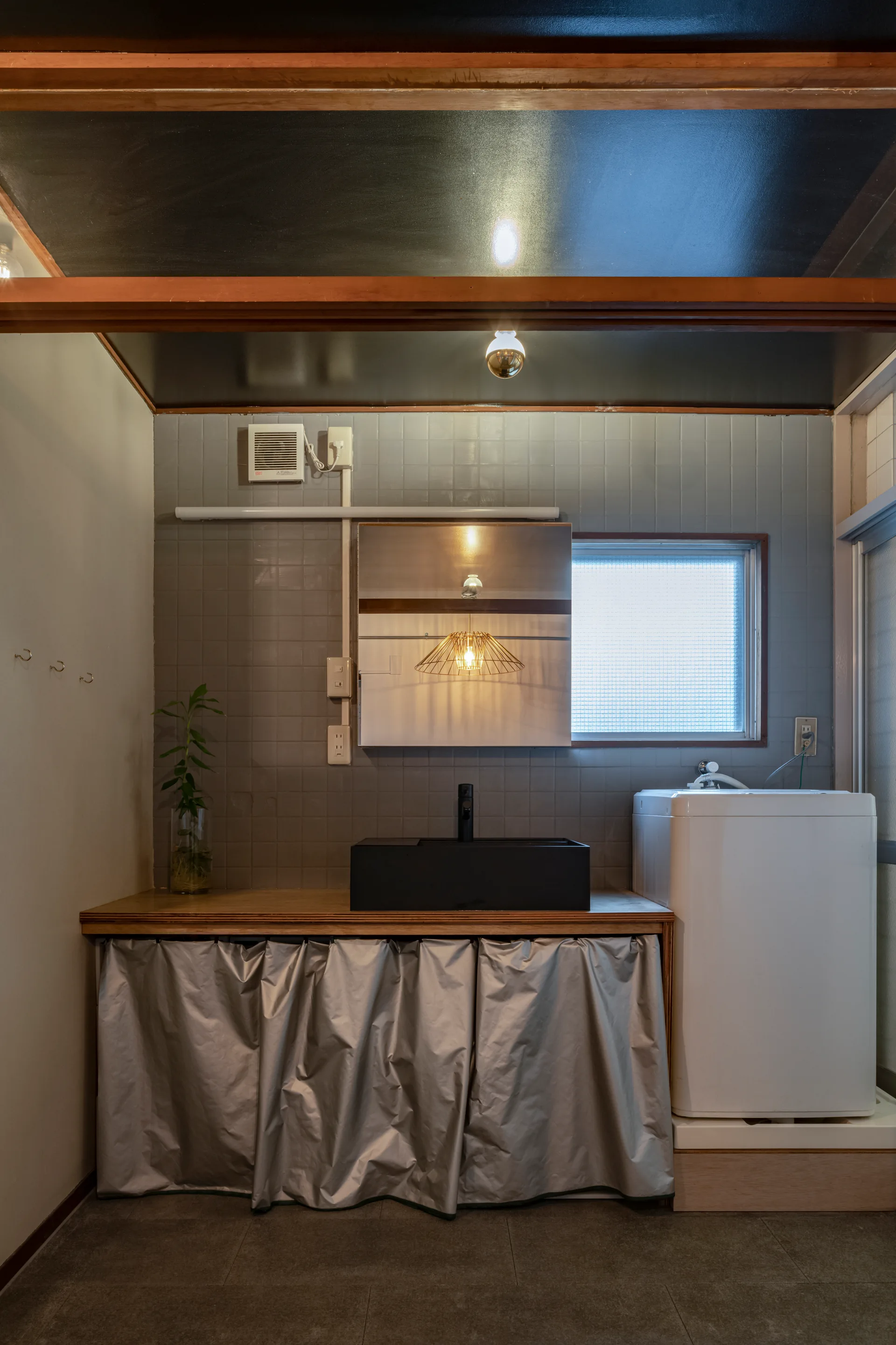 A utility room features a wooden vanity with a black sink and gray fabric skirt, set against gray tiled walls next to a white washing machine.