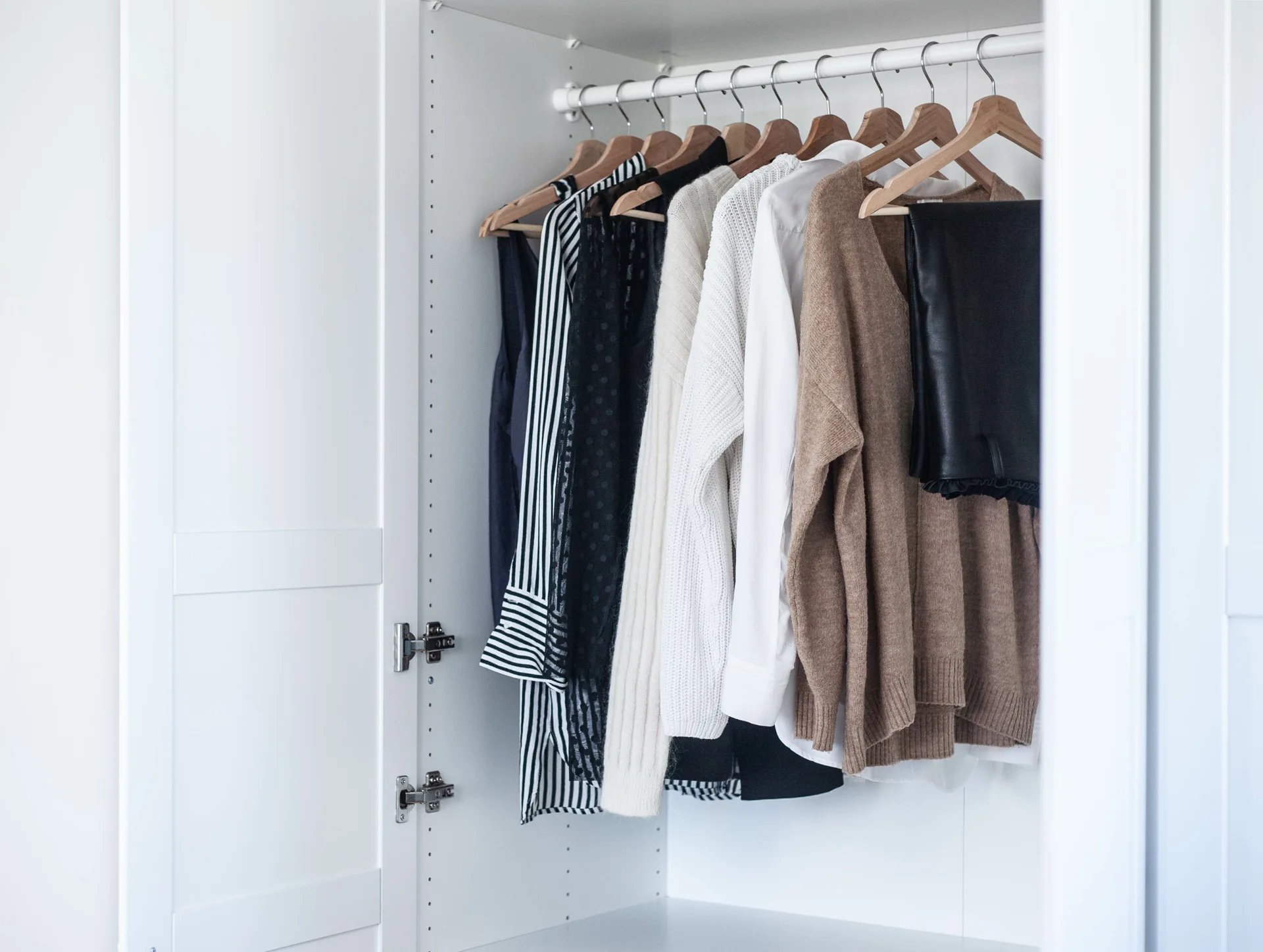 Inside a matte white wardrobe, a row of neutral sweaters and shirts hangs neatly from matching light wood hangers on a white closet rod.