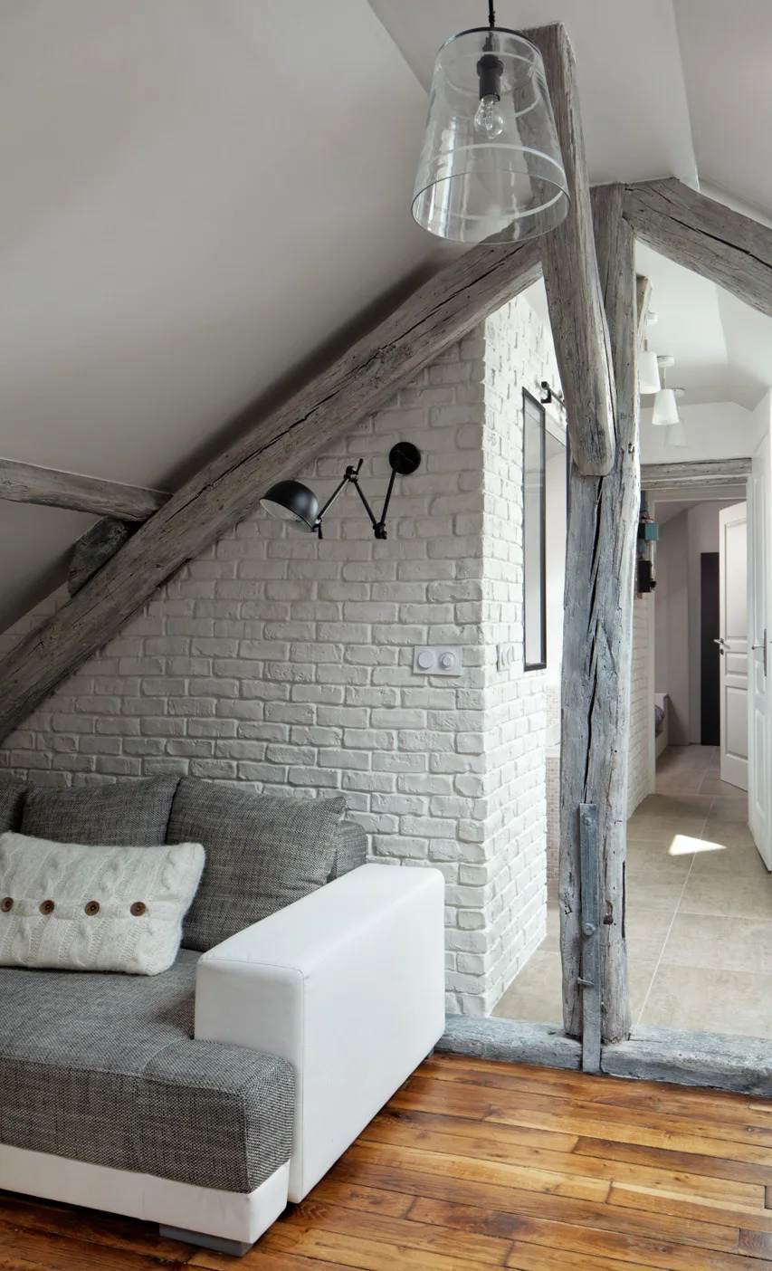 An attic living area features a gray sofa on wood flooring next to a white painted brick wall under sloping ceilings with exposed weathered timber beams.