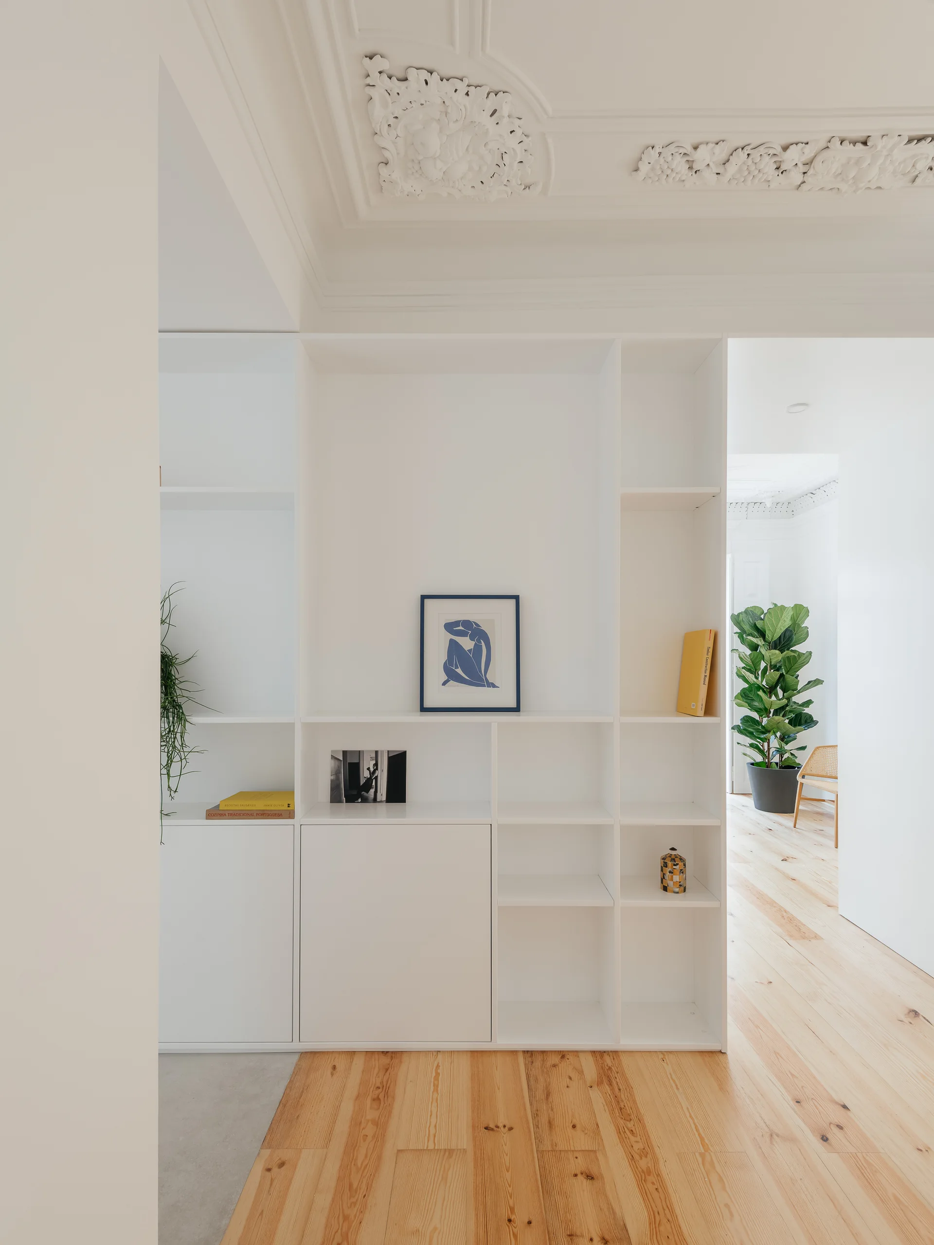 A white floor-to-ceiling storage unit stands under an ornate plaster ceiling in a bright room with light wood floors.