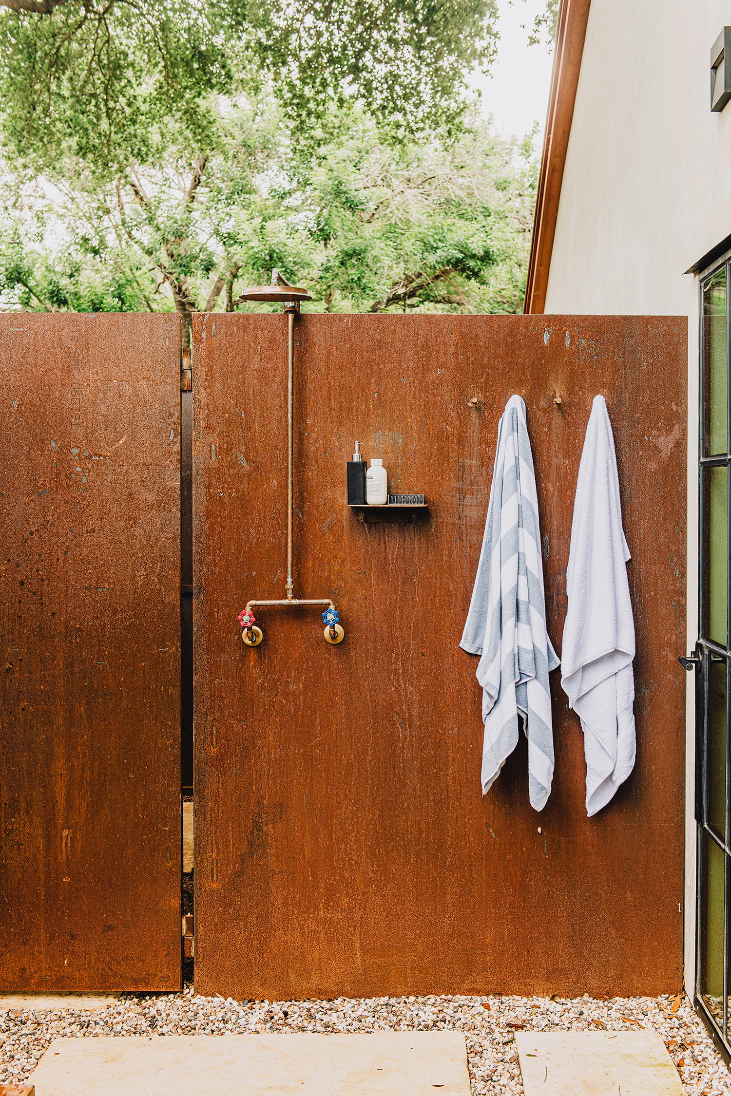 An outdoor brass shower is mounted on a rusted steel privacy wall next to hanging towels and a gravel floor.